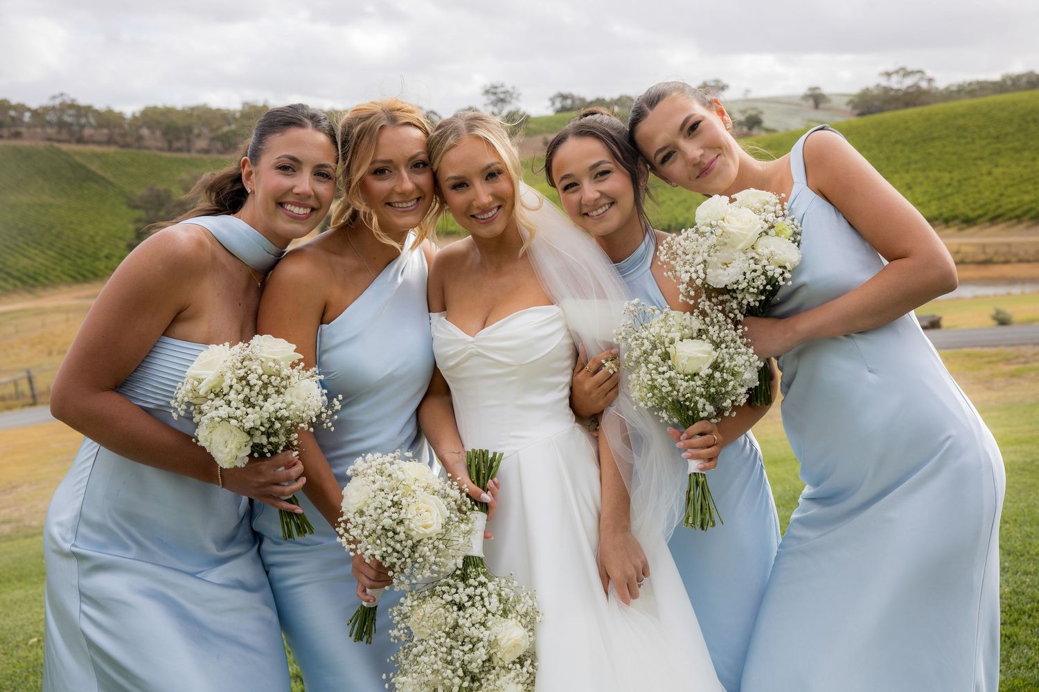 Smiling bride with veil poses with four bridesmaids in light blue dresses holding white bouquets.