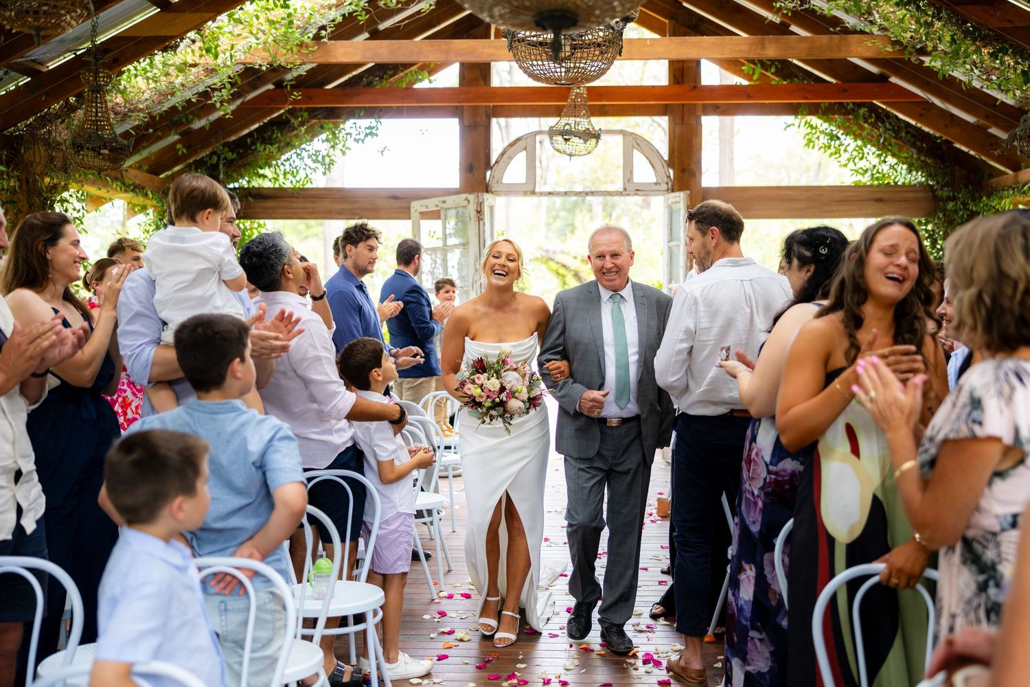 Two brides walk back down the aisle celebrating after their wedding ceremony as guests cheer and toss petals.