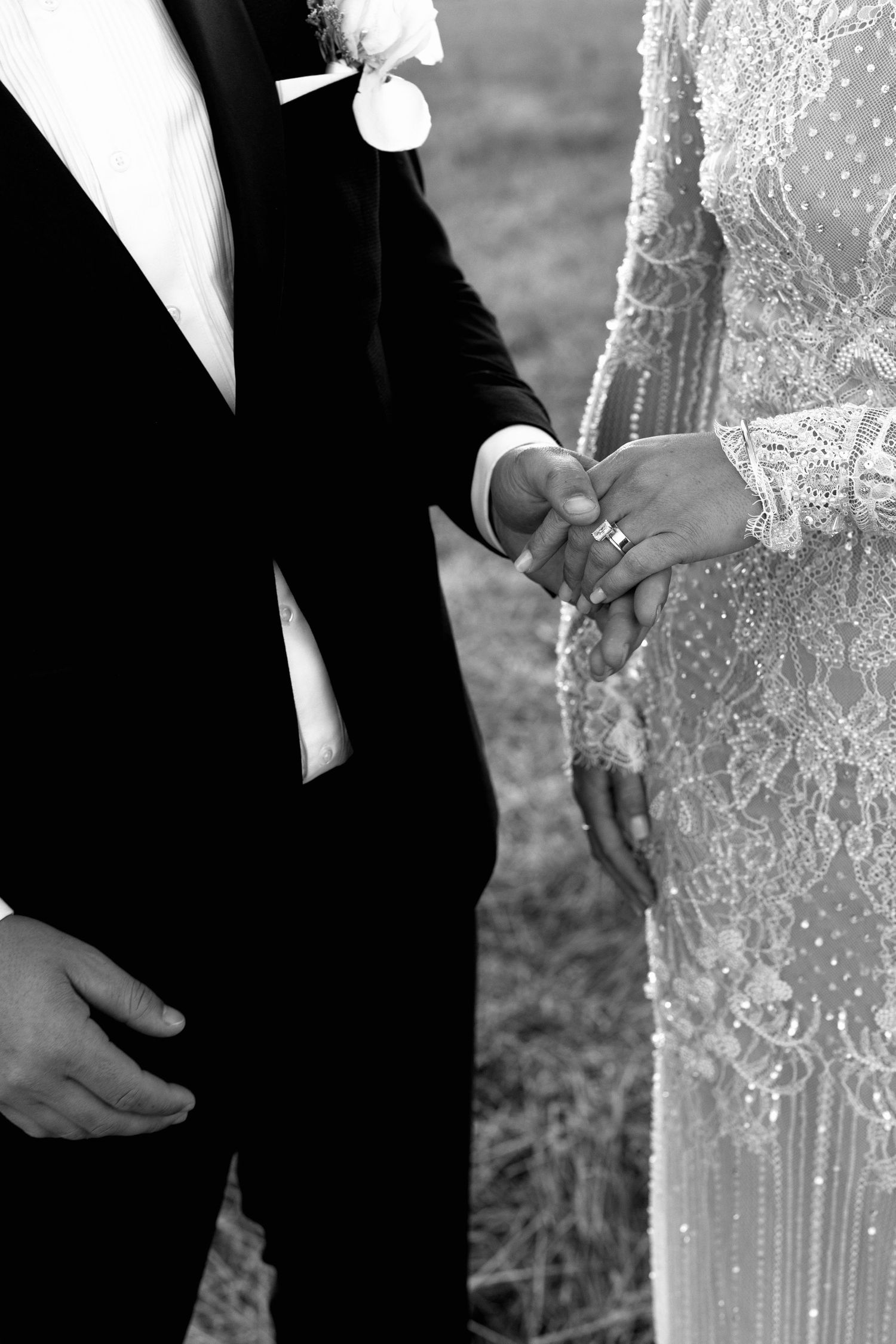 Black and white close-up of bride and groom holding hands, showcasing wedding rings and beaded bridal gown detail.