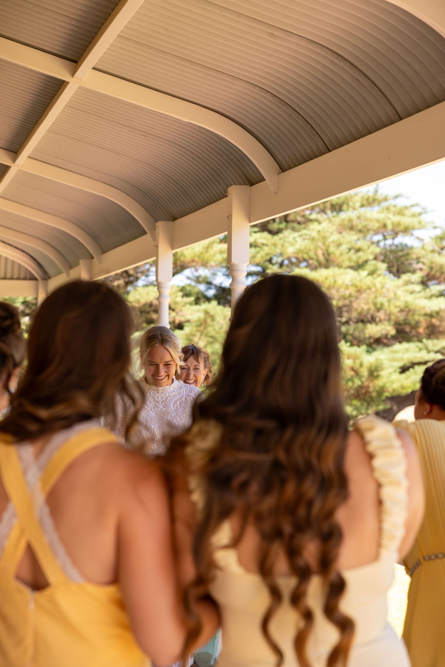 Bride smiles as bridesmaids in yellow dresses gather around her under a covered outdoor area.