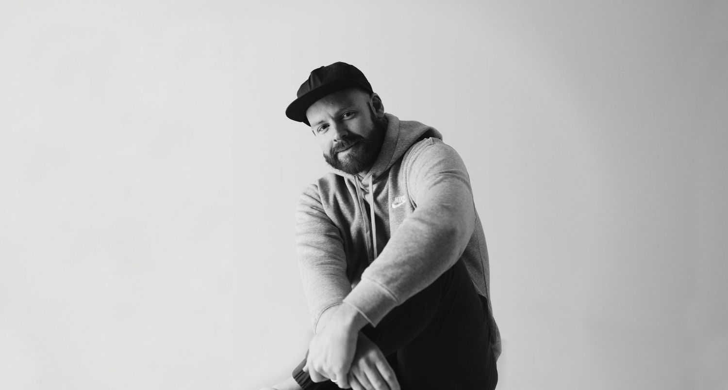 Black and white portrait of a bearded man wearing a cap and hoodie, leaning forward in a relaxed pose.