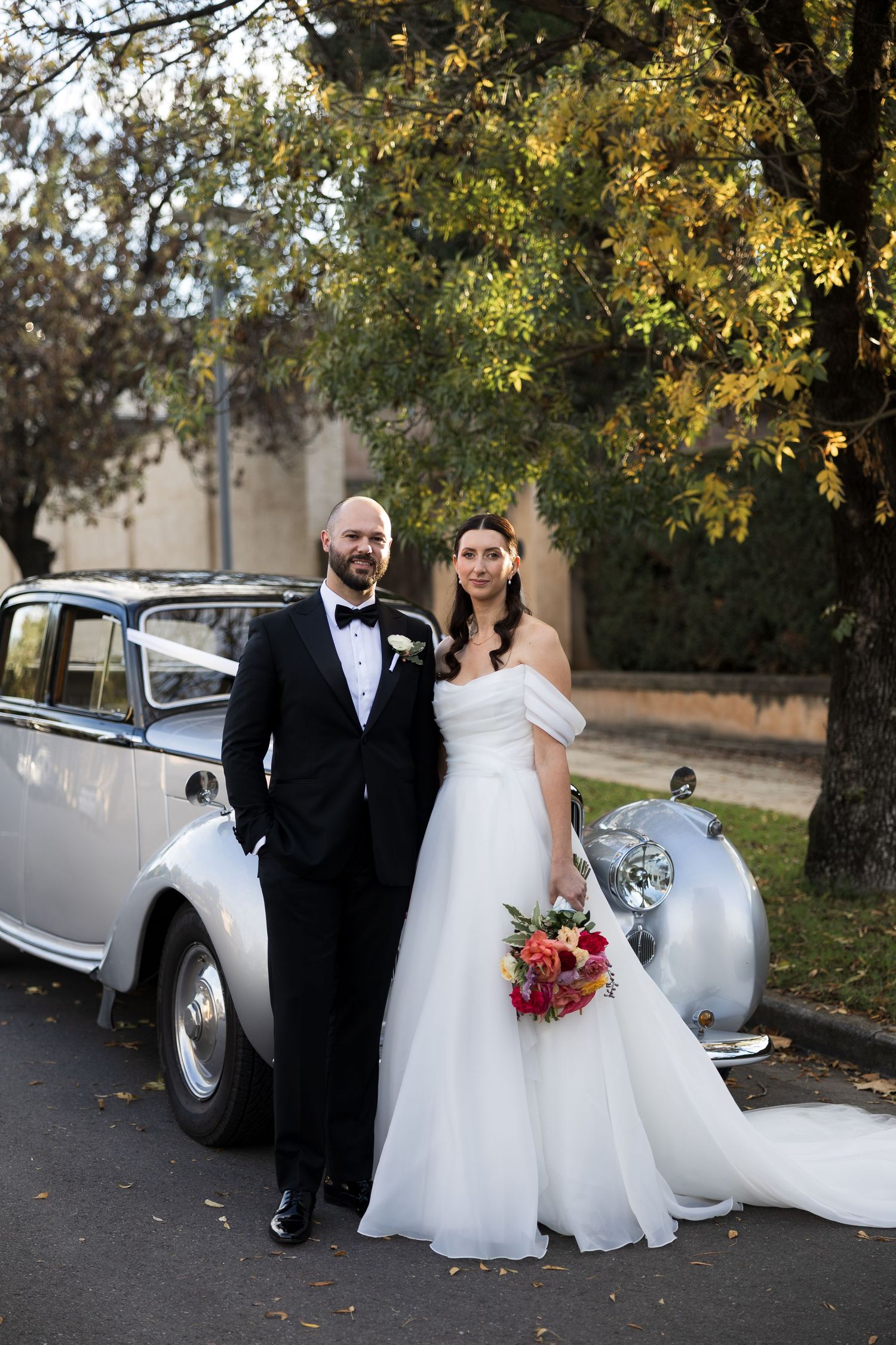 Bride and groom posing beside a vintage silver car, she holding a colorful bouquet in an elegant white dress.