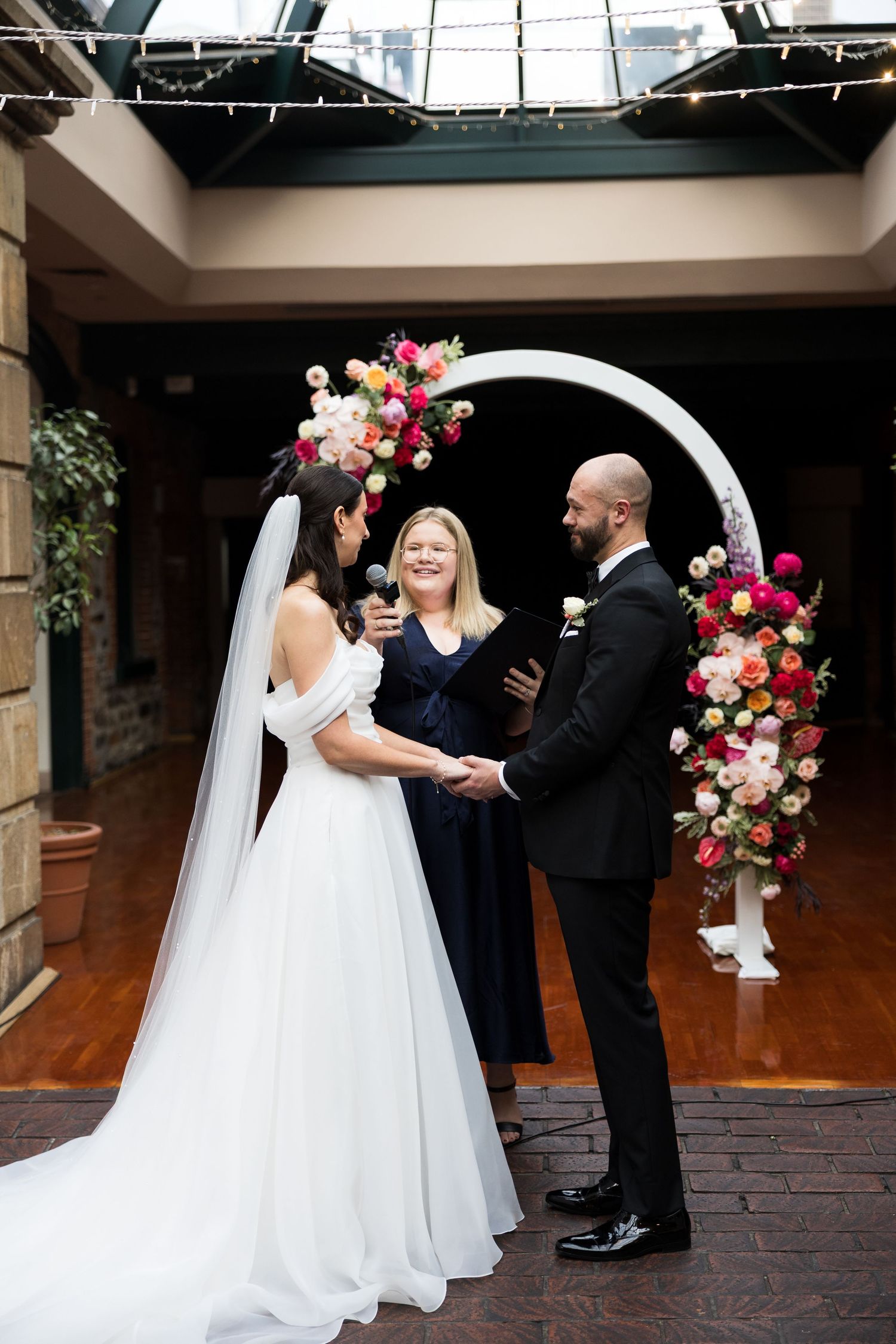 Bride and groom hold hands at indoor wedding ceremony before a circular arch adorned with vibrant pink and red floral arrangements.