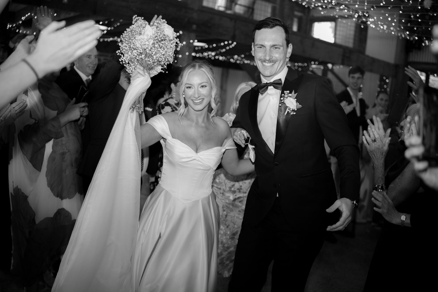 Joyful bride and groom exit wedding reception amid cheering guests in black and white photo.