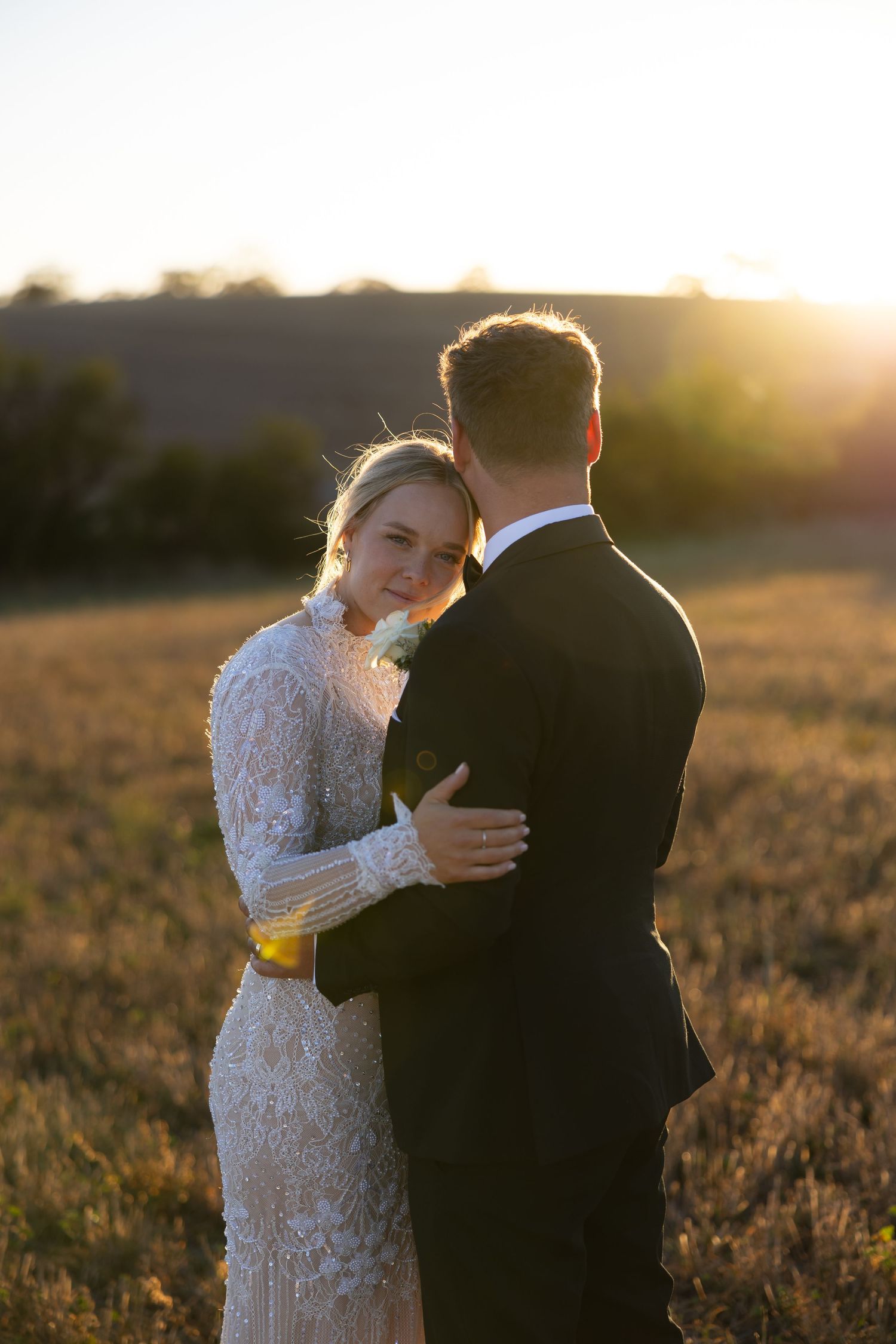 Bride in lace long-sleeve gown embracing groom in a golden sunset field.