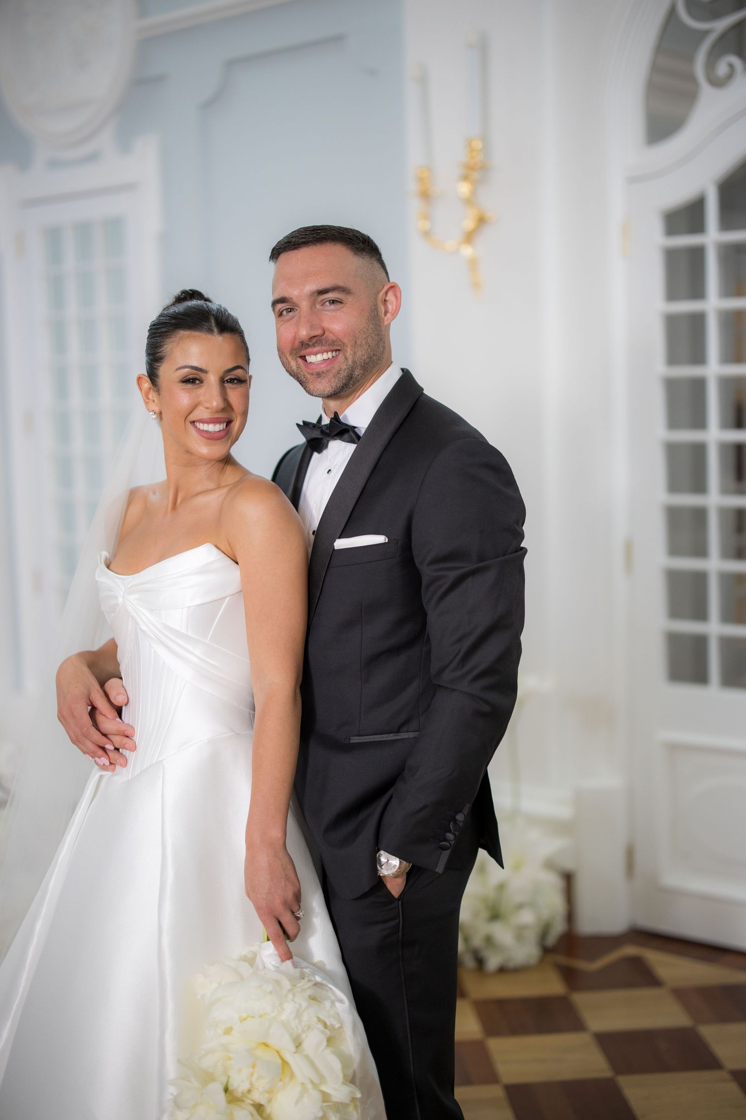 Happy couple on wedding day, bride holding white bouquet in satin gown, groom in black bow tie tuxedo.