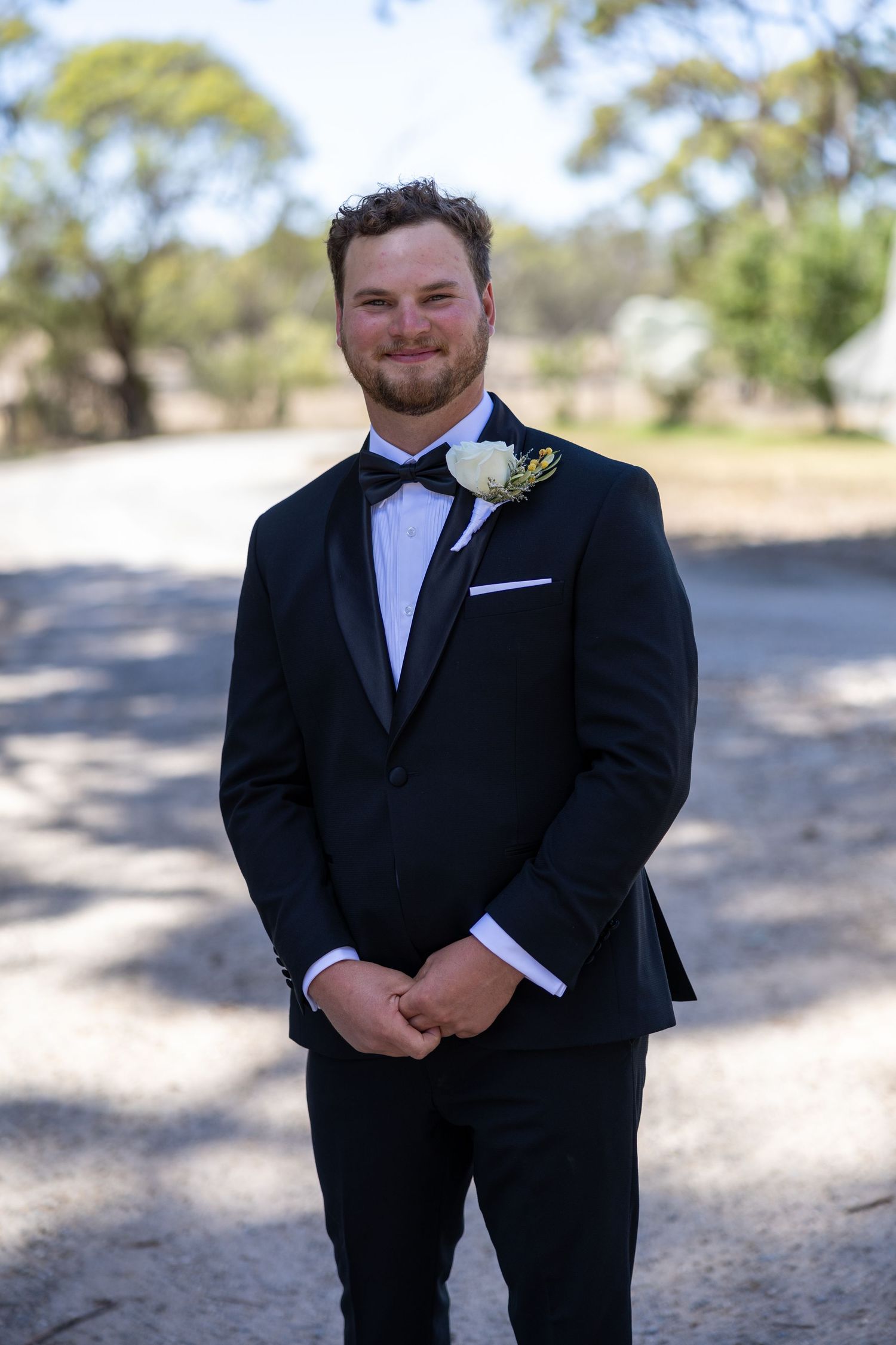 Smiling groom in black tuxedo with bow tie and white boutonniere standing outdoors.