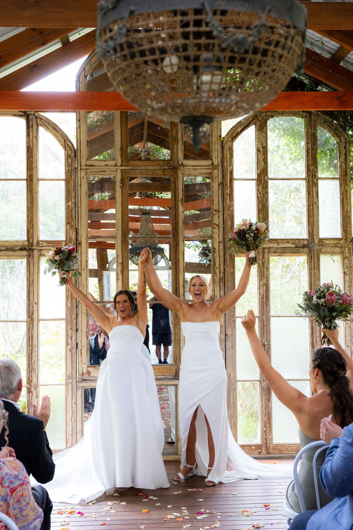 Two brides raise their bouquets triumphantly after exchanging vows in a stunning rustic wedding chapel.