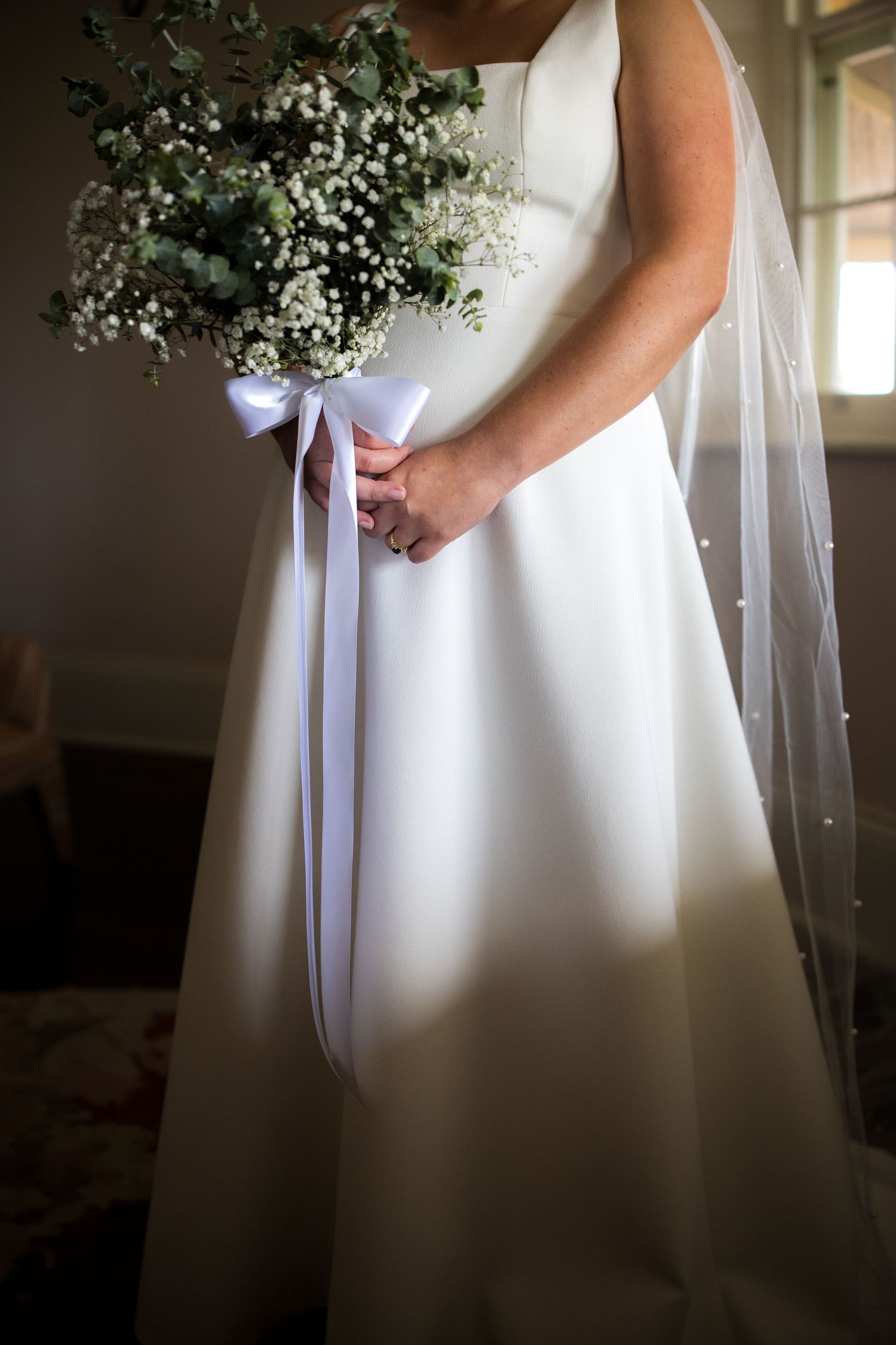 Bride in white satin gown holds a baby's breath and eucalyptus bouquet with ribbon bow.