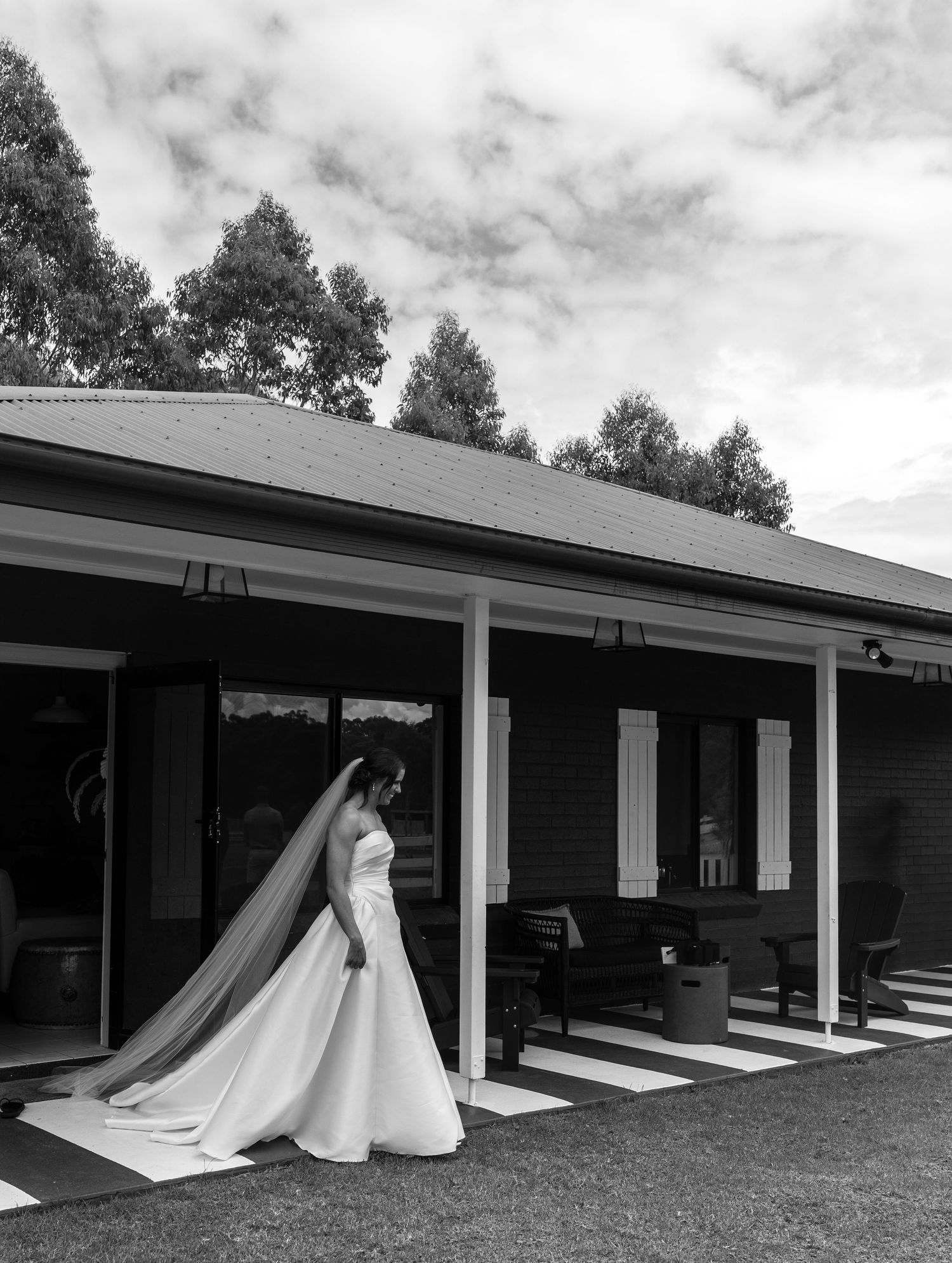 Black and white photo of bride in white gown with long veil standing outside a dark wooden building.