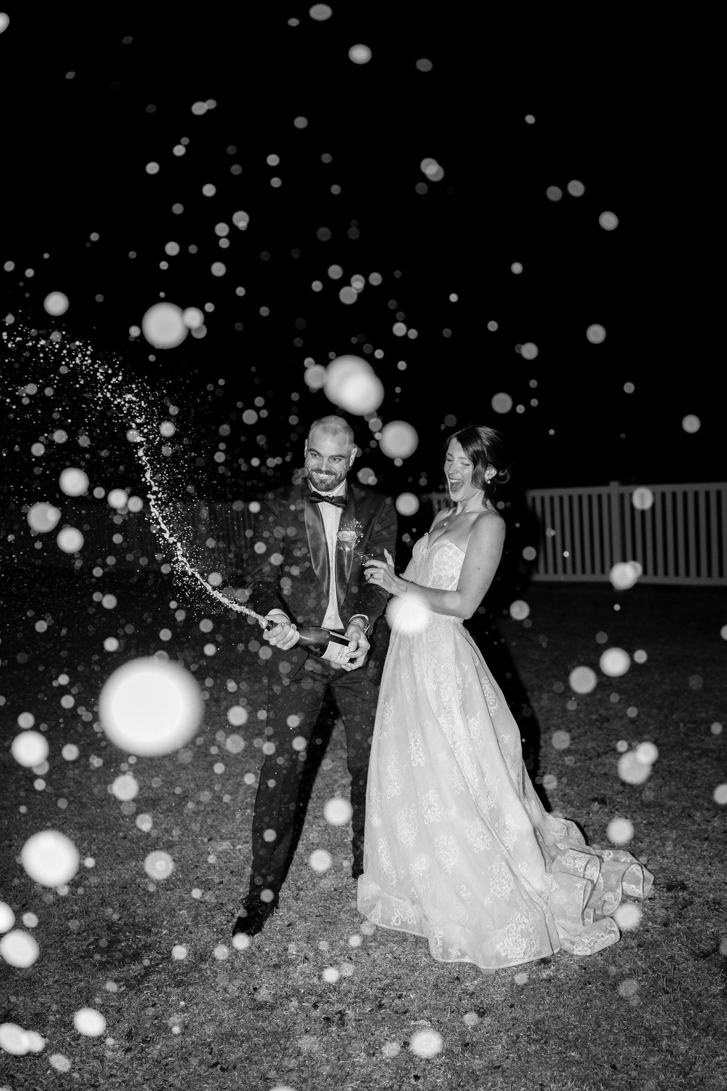 Black and white photo of newlyweds celebrating with champagne spray at nighttime wedding reception.