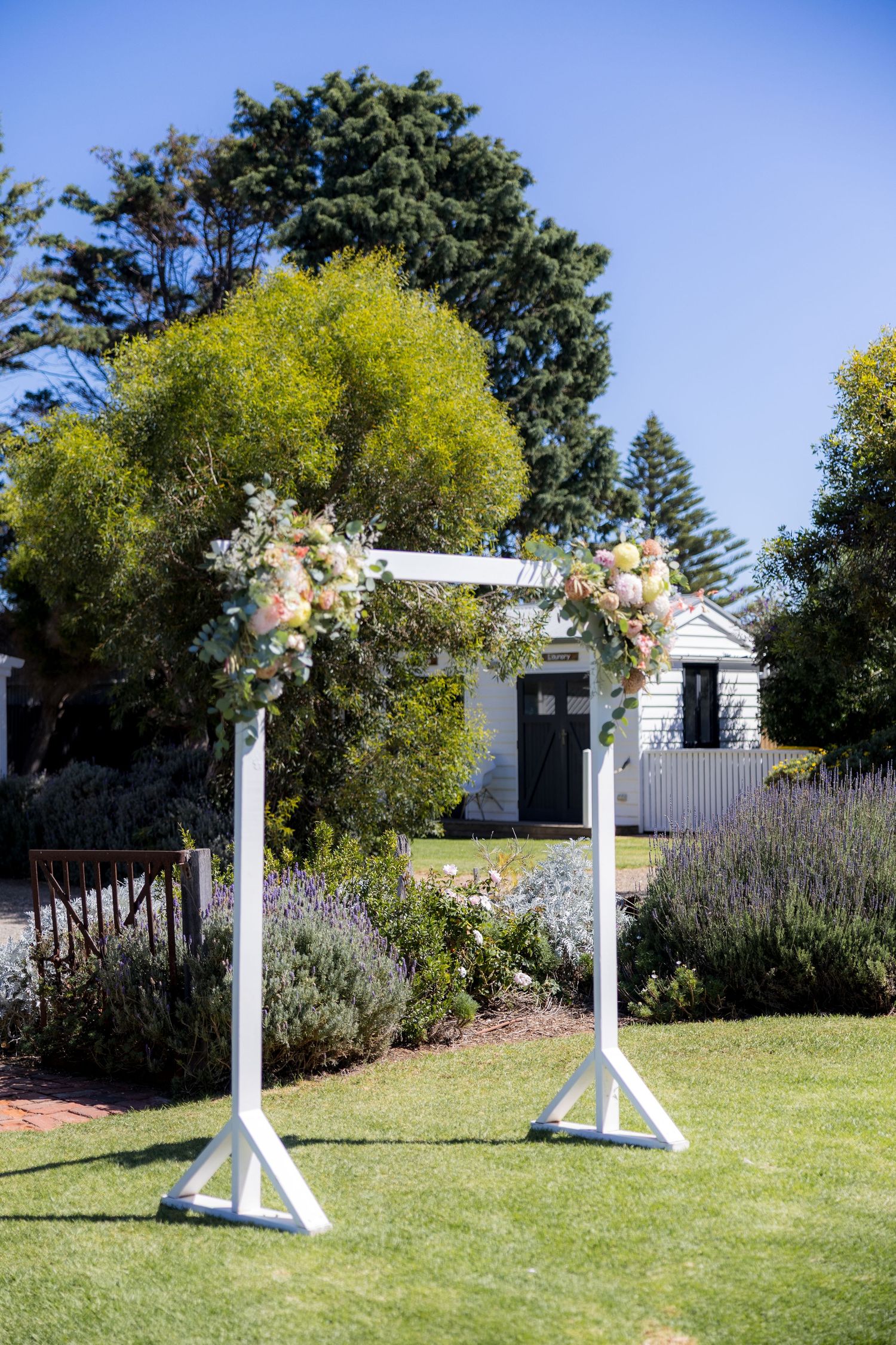 White wedding arch adorned with pink and white flowers in a lush garden with a white cottage backdrop.
