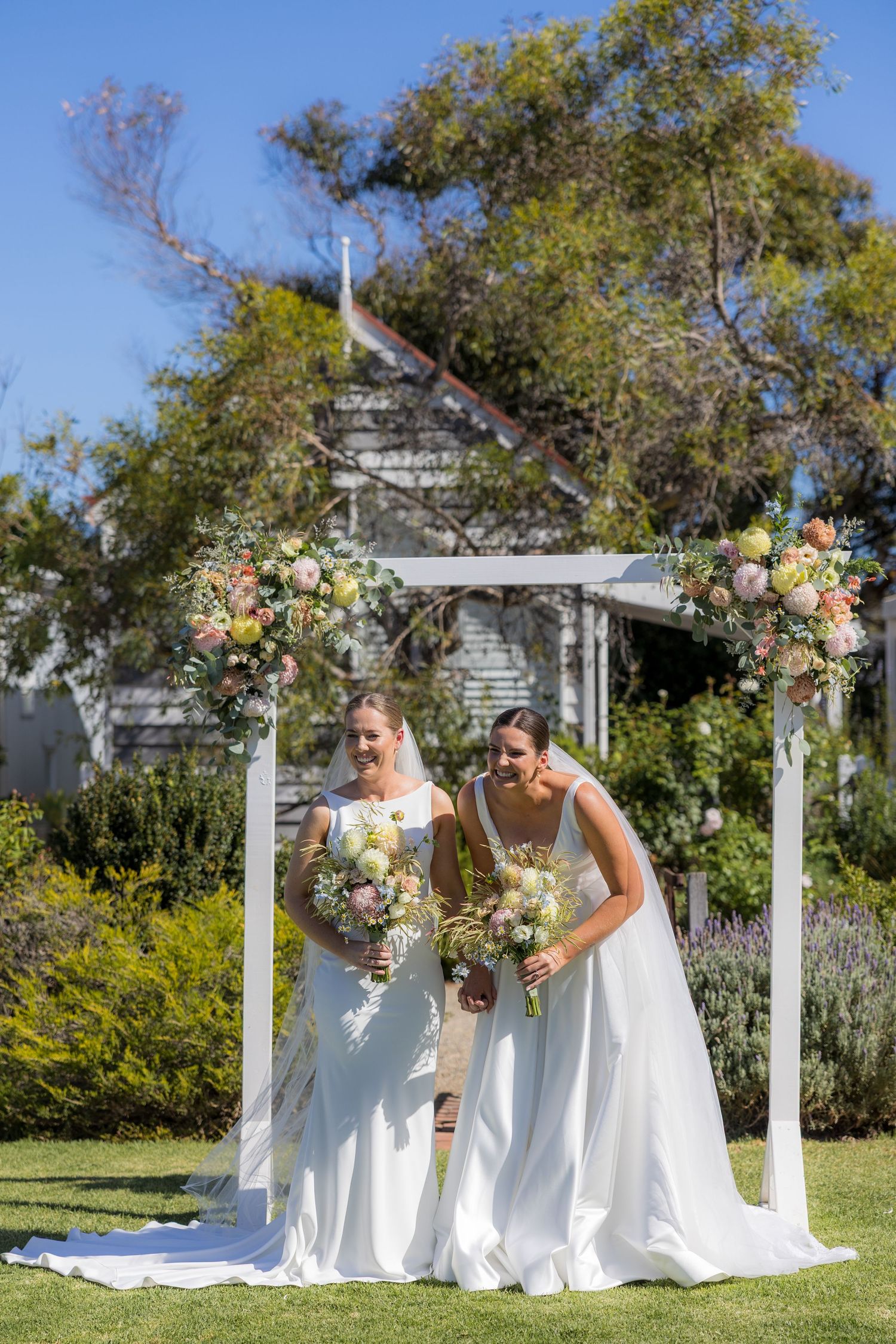Two brides in white gowns holding bouquets stand under a floral arch in a garden wedding ceremony.