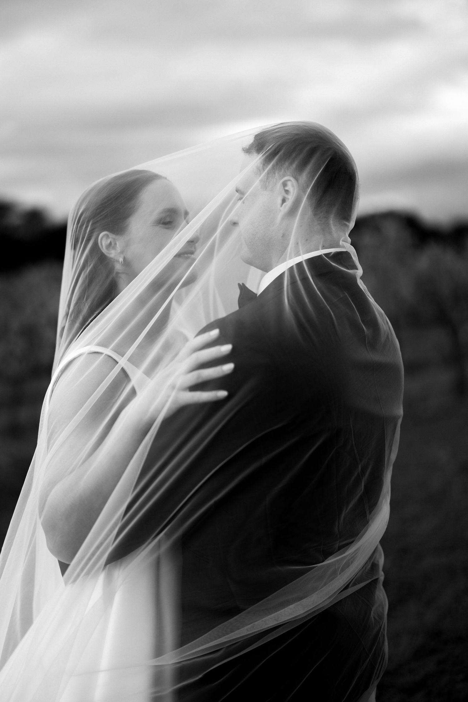 Black and white photo of bride and groom embracing under flowing wedding veil outdoors.