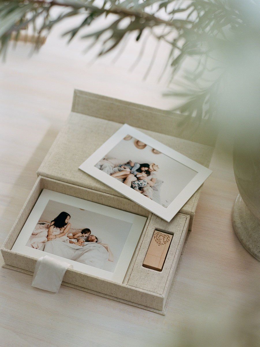 Open linen photo keepsake box with printed photos and engraved USB drive on wooden surface with greenery.