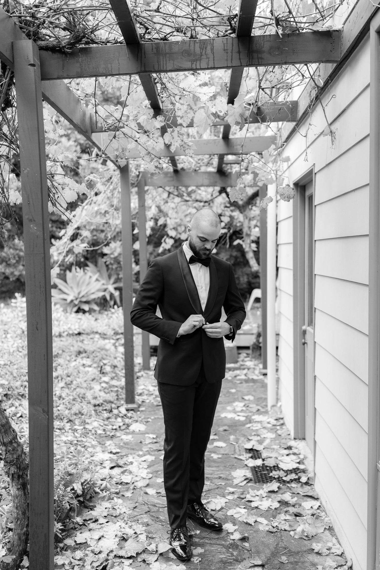Dapper groom in black tuxedo adjusting jacket under rustic pergola surrounded by autumn leaves.