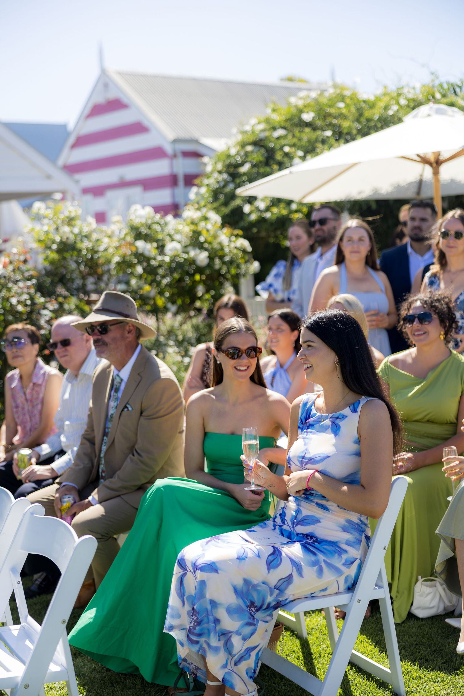 Smiling wedding guests seated outdoors in white chairs enjoying a sunny garden ceremony.