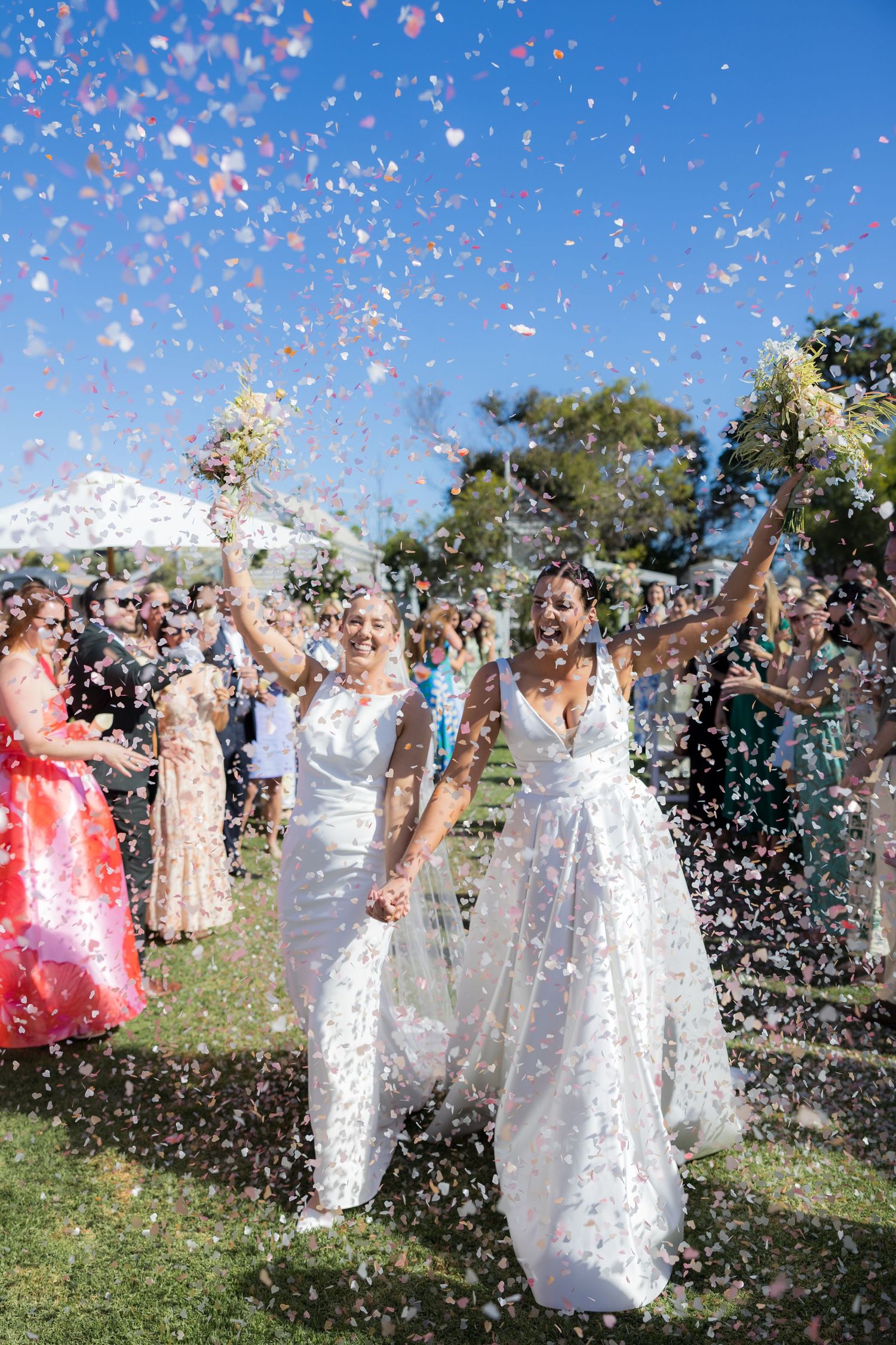 Two brides celebrate joyfully as colorful confetti fills the air during their outdoor wedding reception.