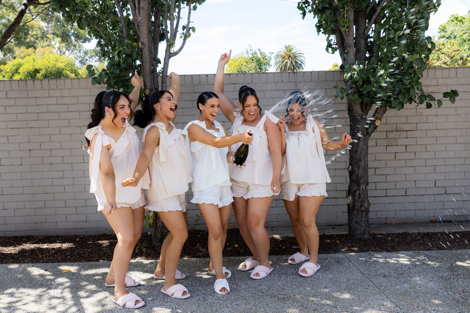 Five bridesmaids in white pajamas celebrating with champagne outdoors before the wedding.