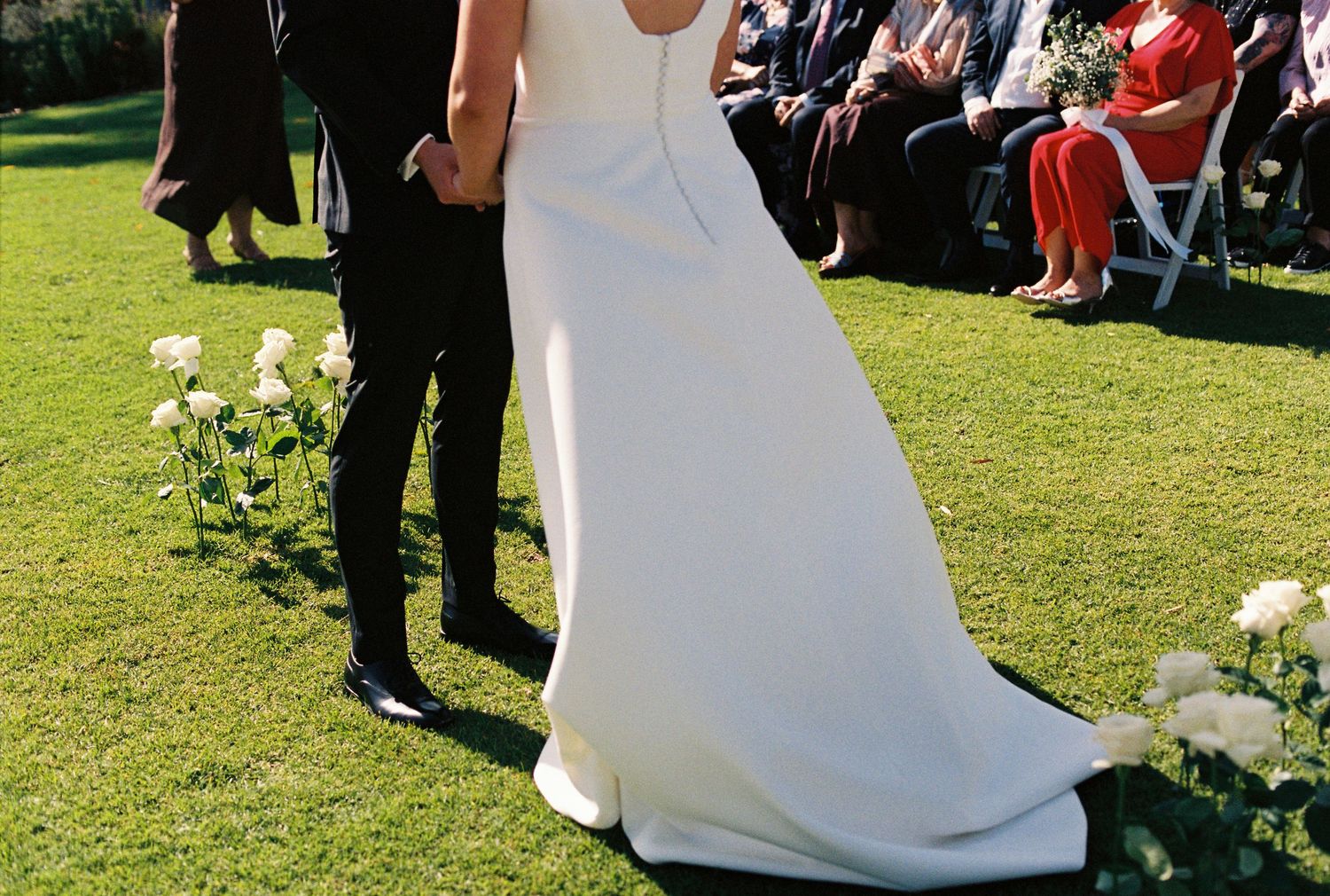 Bride in elegant white gown holds hands with groom in black suit during outdoor wedding ceremony on green lawn.