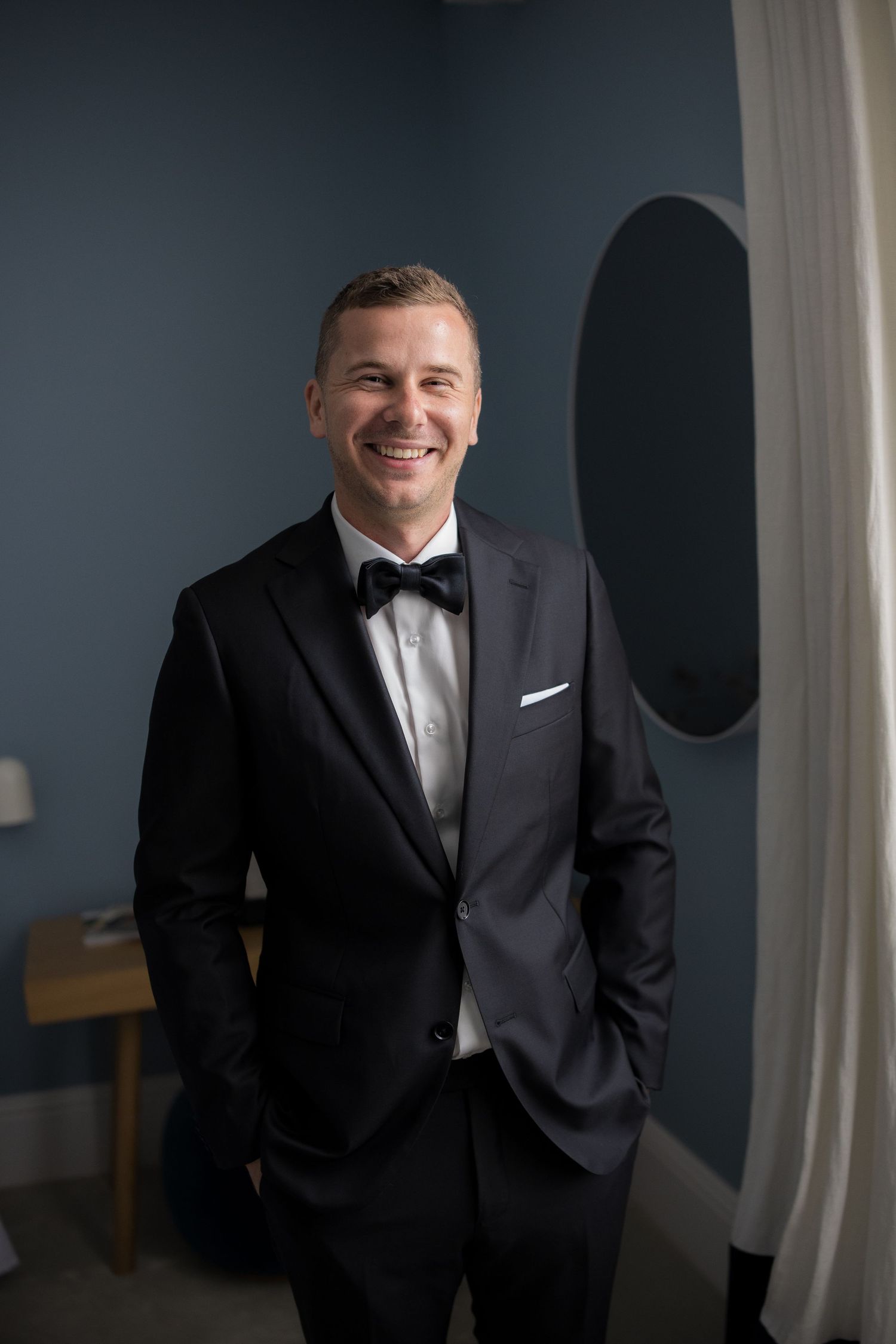 Smiling groom in black tuxedo with bow tie poses confidently in a blue-walled room.