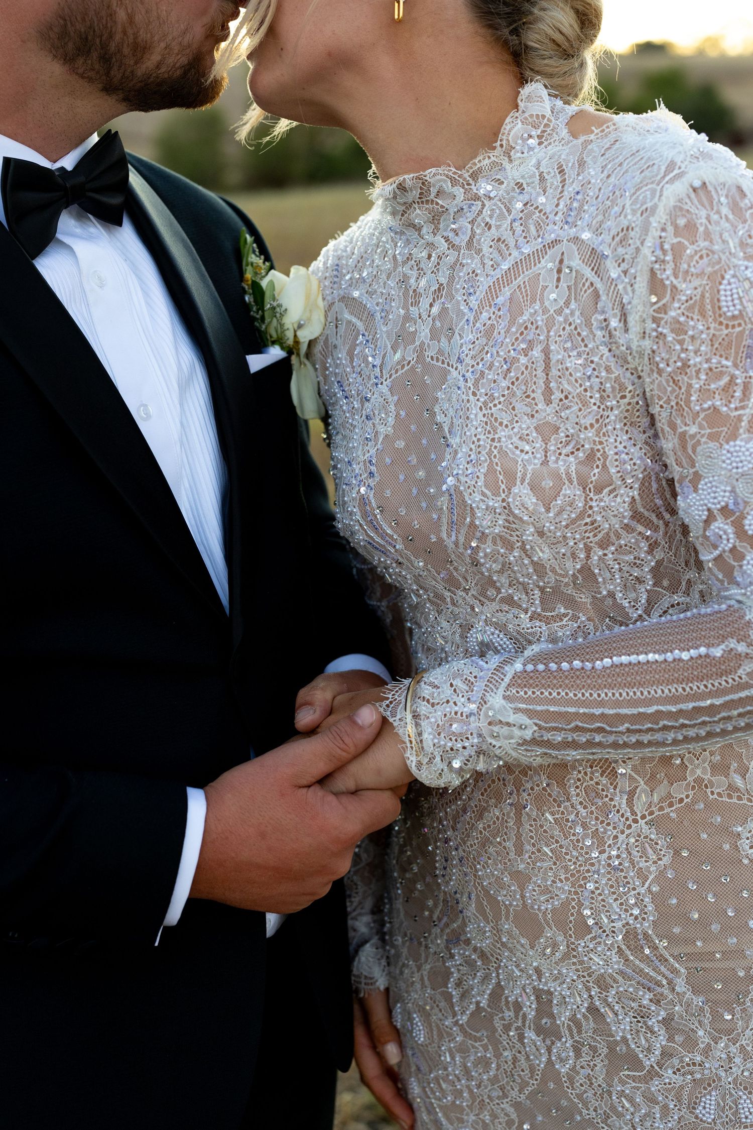 Close-up of bride in intricate lace long-sleeve wedding gown holding hands with groom in black tuxedo.