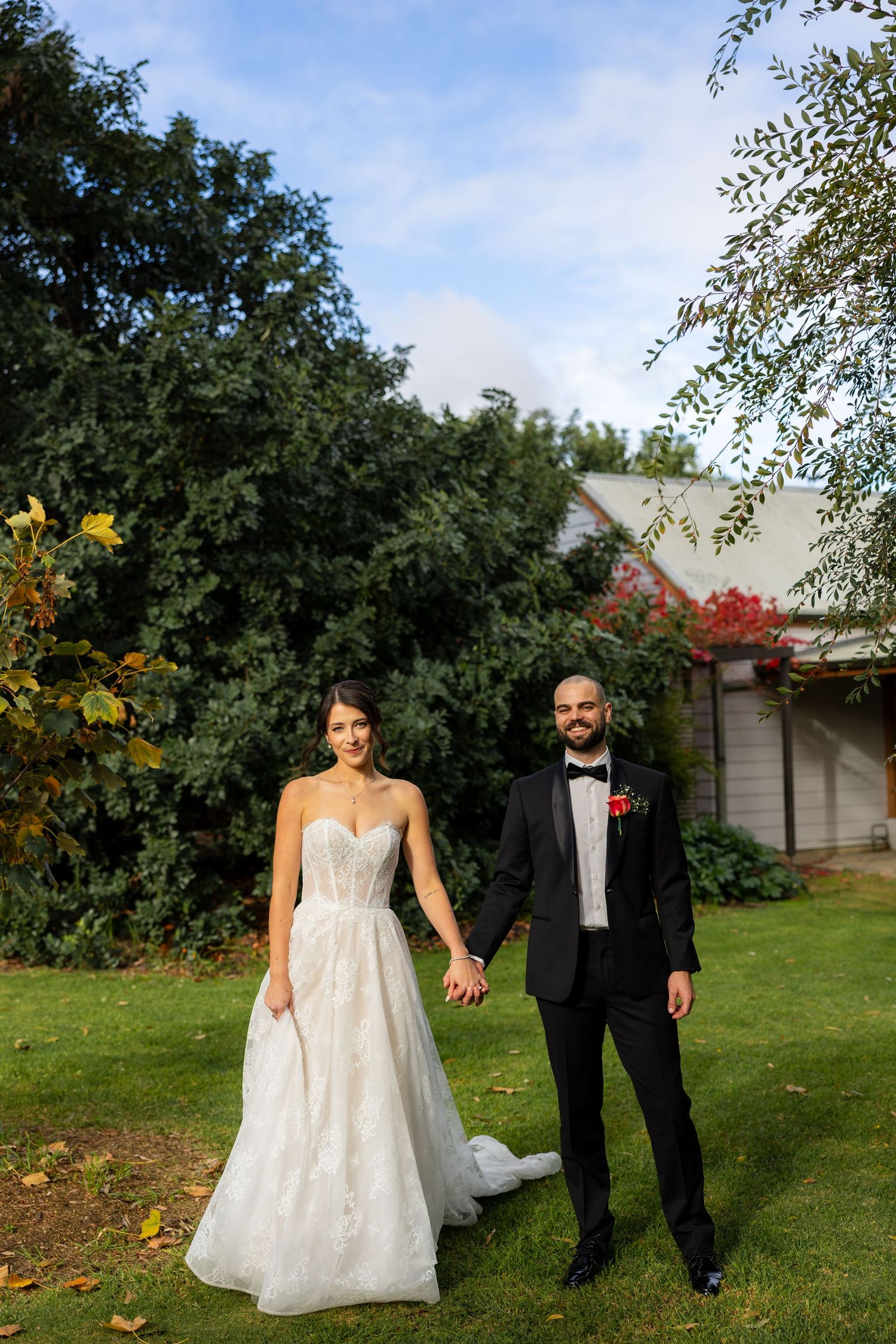 Smiling bride and bearded groom hold hands on a lush green lawn outside a charming wedding venue.