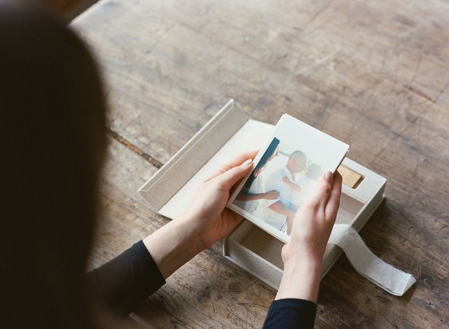 Person opening a white photo keepsake box with ribbon on a wooden table revealing printed photos inside.