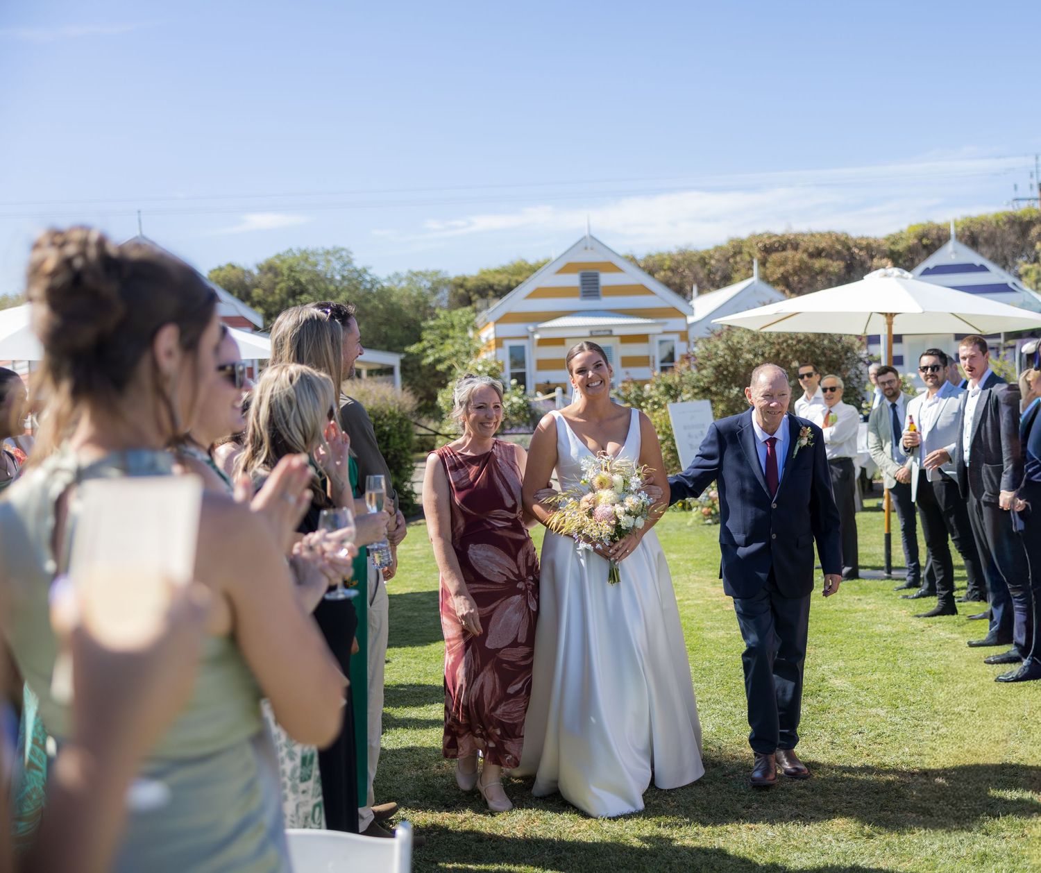Bride escorted by parents down grassy outdoor aisle with guests watching at seaside wedding.