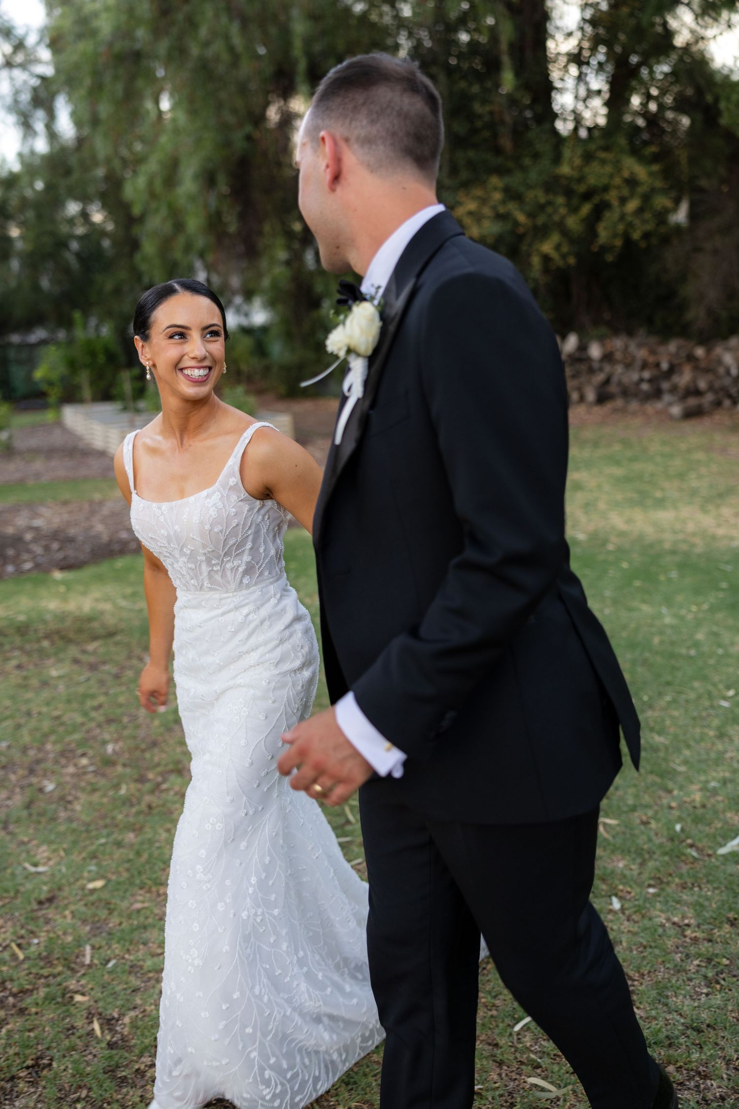 Bride in white lace gown smiles at groom in black tuxedo during outdoor first look.