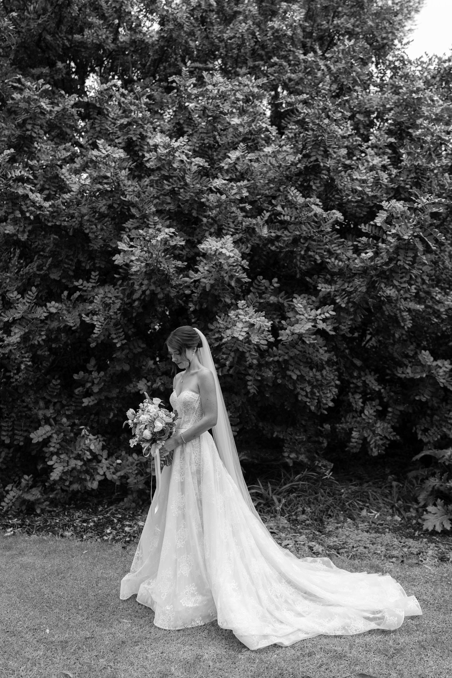 Bride in full lace wedding gown with veil and bouquet stands before lush garden trees.