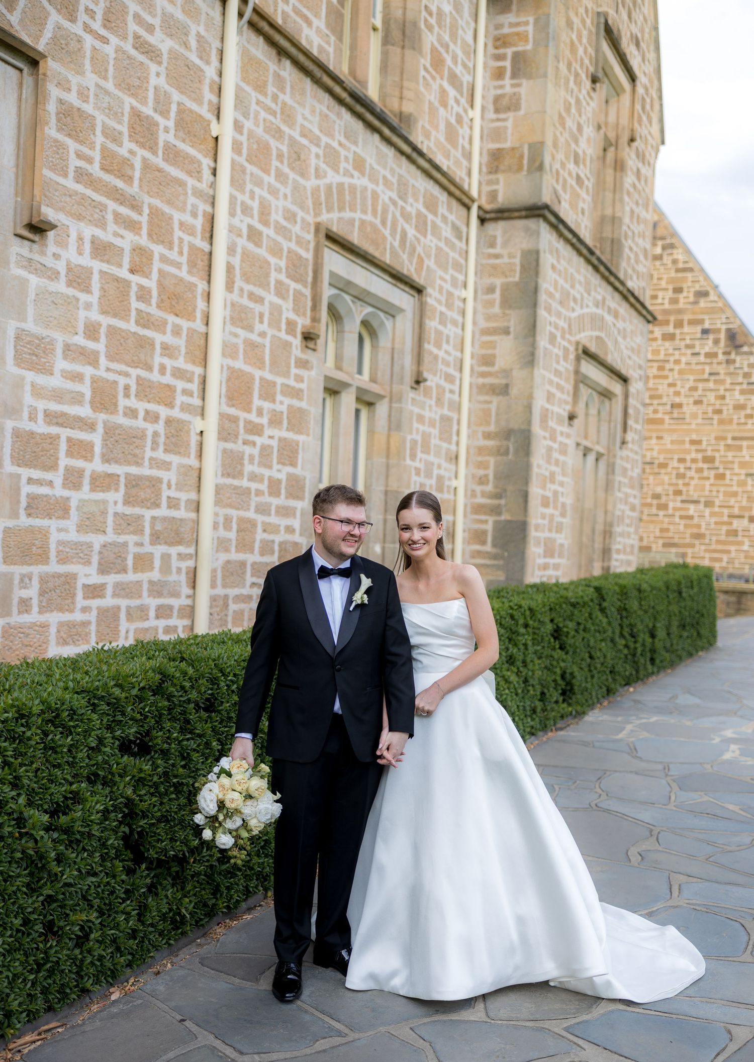 Bride and groom walking hand in hand beside a historic sandstone building on their wedding day.