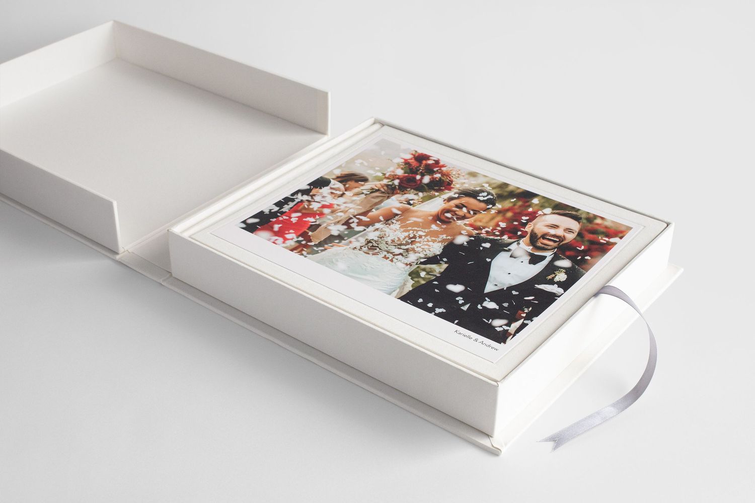 Elegant white wedding photo album box with ribbon, open to reveal a colorful wedding photo inside.