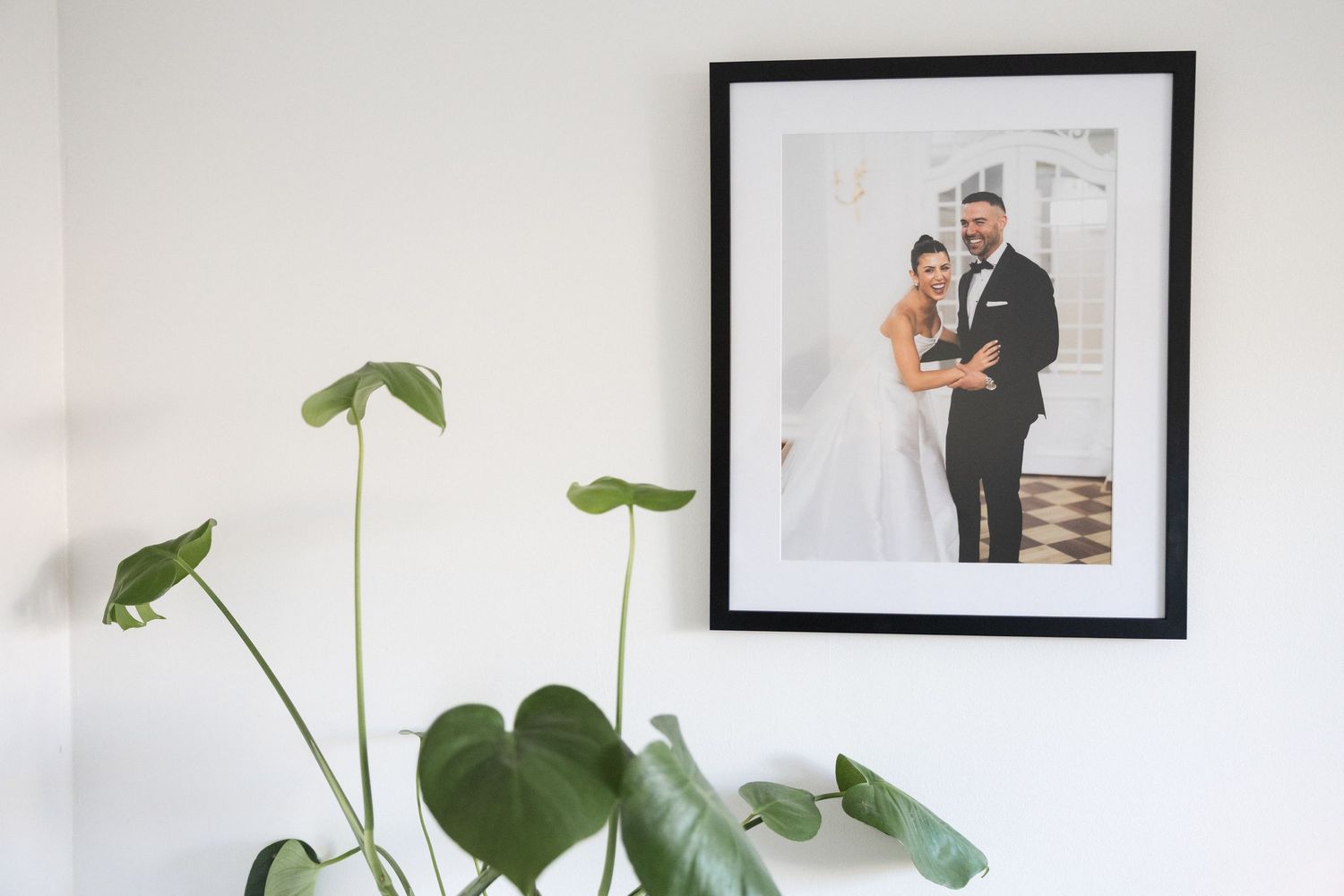 Framed wedding couple portrait on white wall with large monstera plant leaves in foreground.