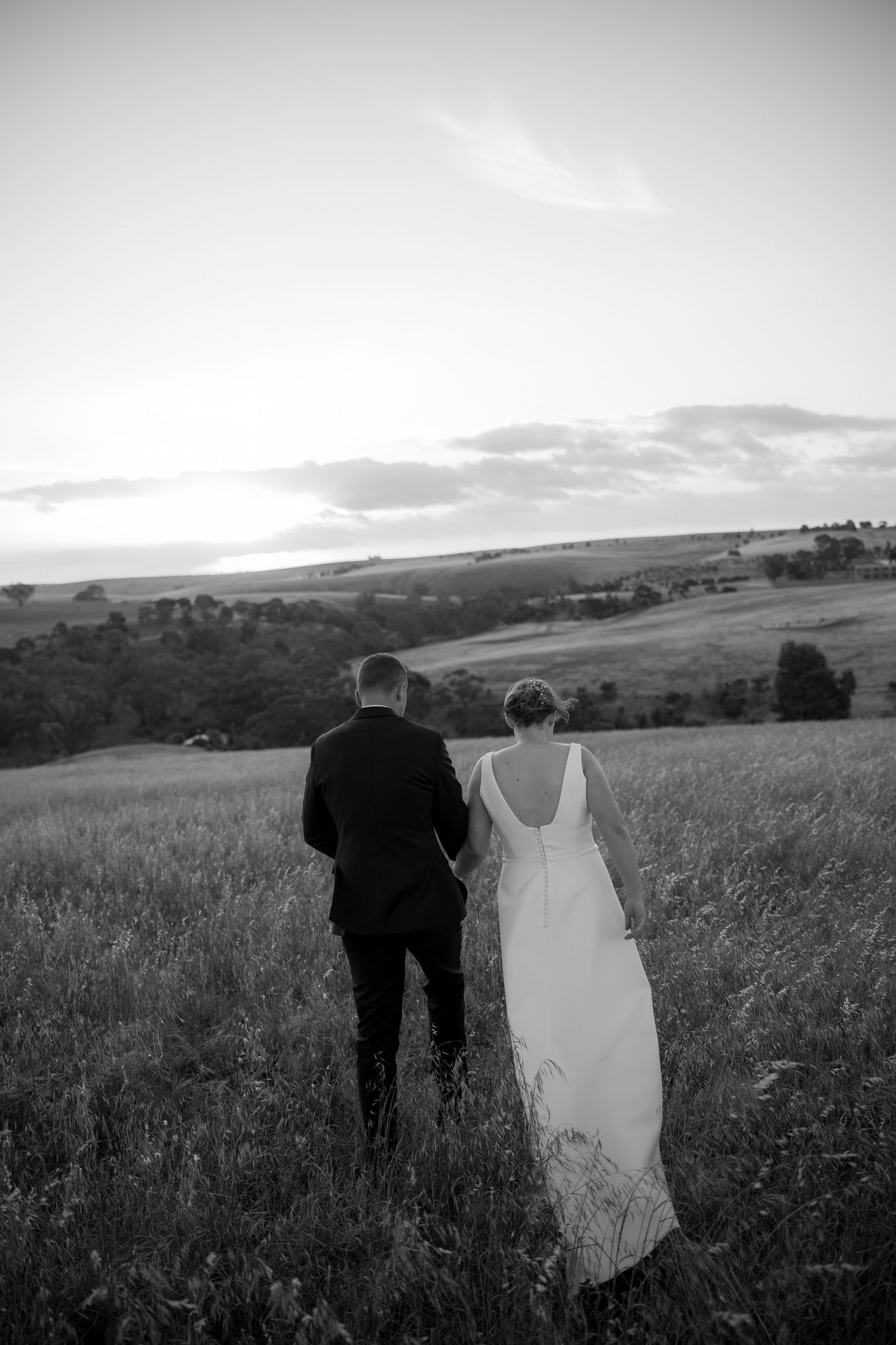 Black and white photo of newlyweds walking hand in hand through open countryside field.