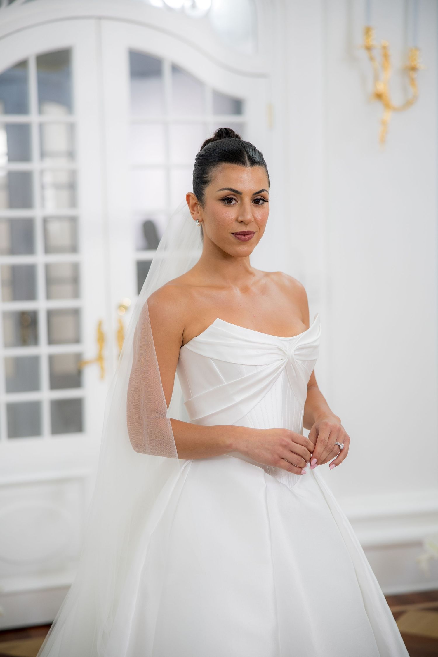 Elegant bride in a strapless white satin ballgown with veil posing in a bright white ornate ballroom.