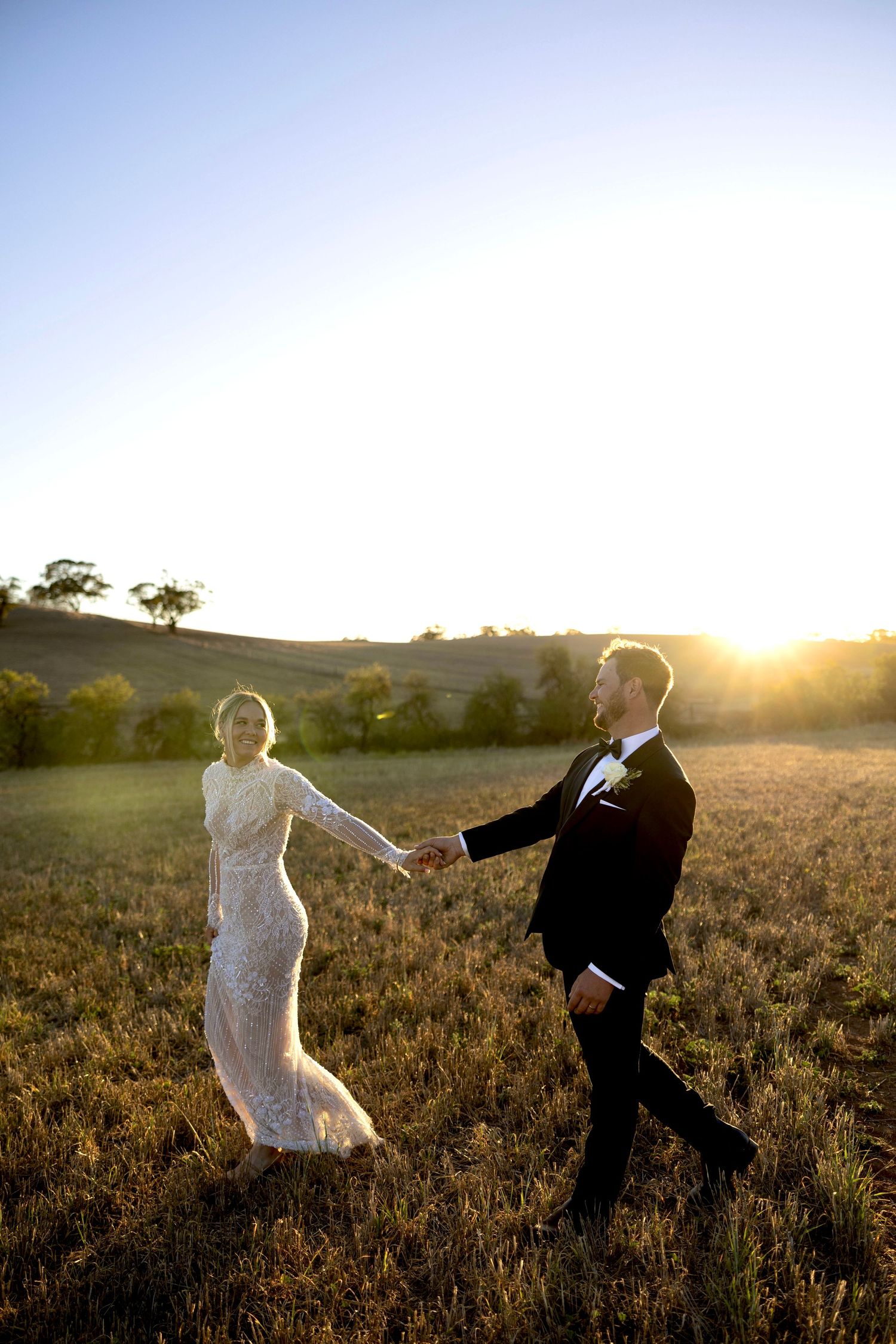 Bride and groom holding hands walking through a golden field at sunset during their outdoor wedding celebration.