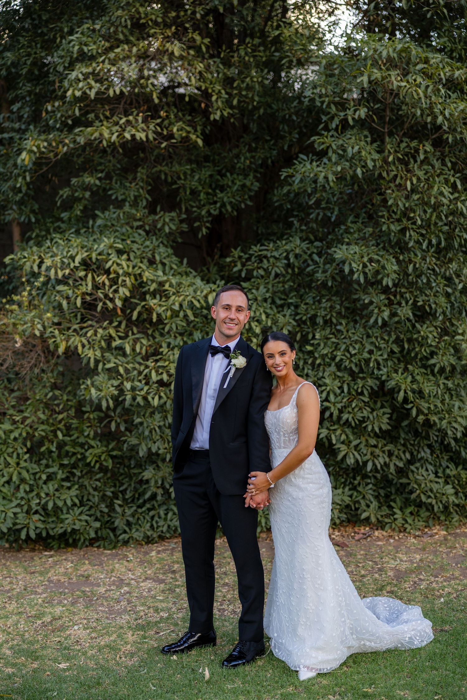 Bride and groom posing together in garden, she in fitted lace gown, he in black tuxedo with bow tie.