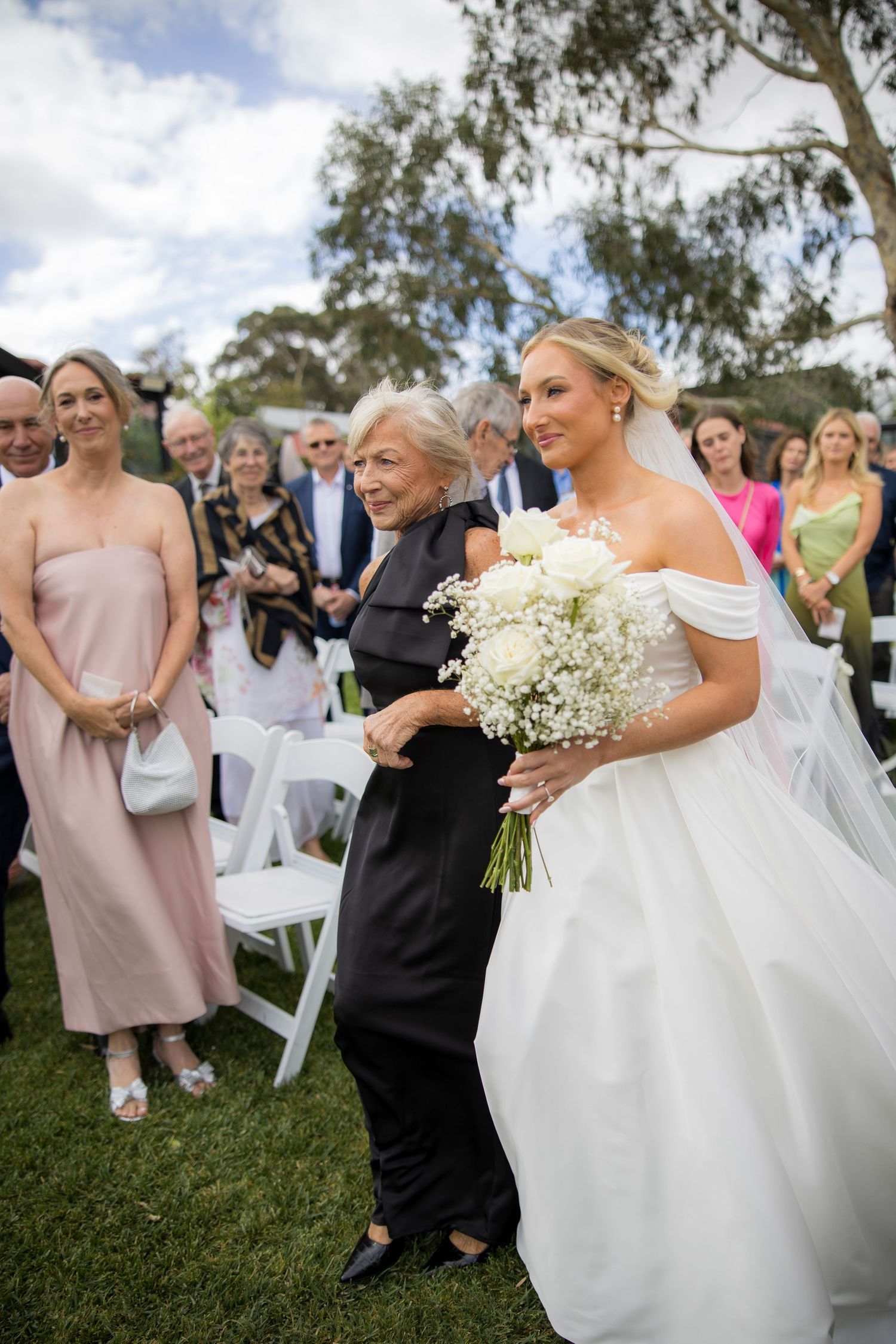 Bride walking down the aisle in white off-shoulder gown escorted by elderly family member.