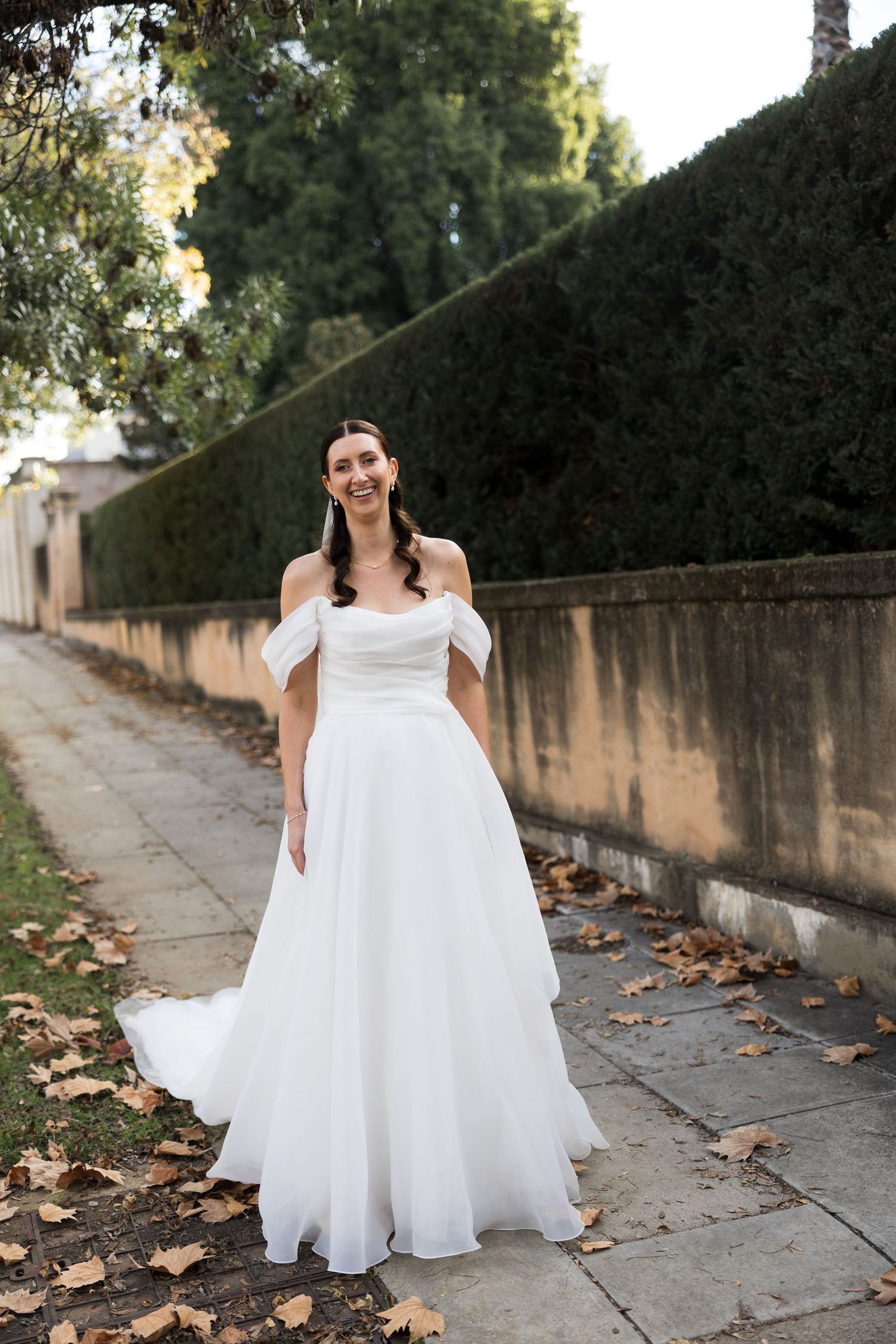 Smiling bride in off-shoulder white ball gown wedding dress standing on autumn leaf-covered path beside stone wall.