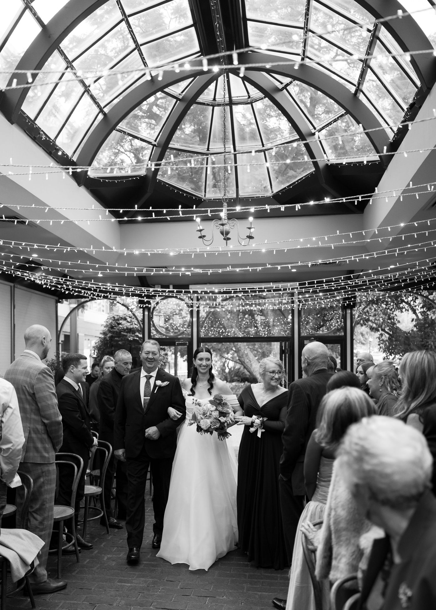 Bride walks down the aisle in a glass-domed venue adorned with string lights during an elegant wedding ceremony.