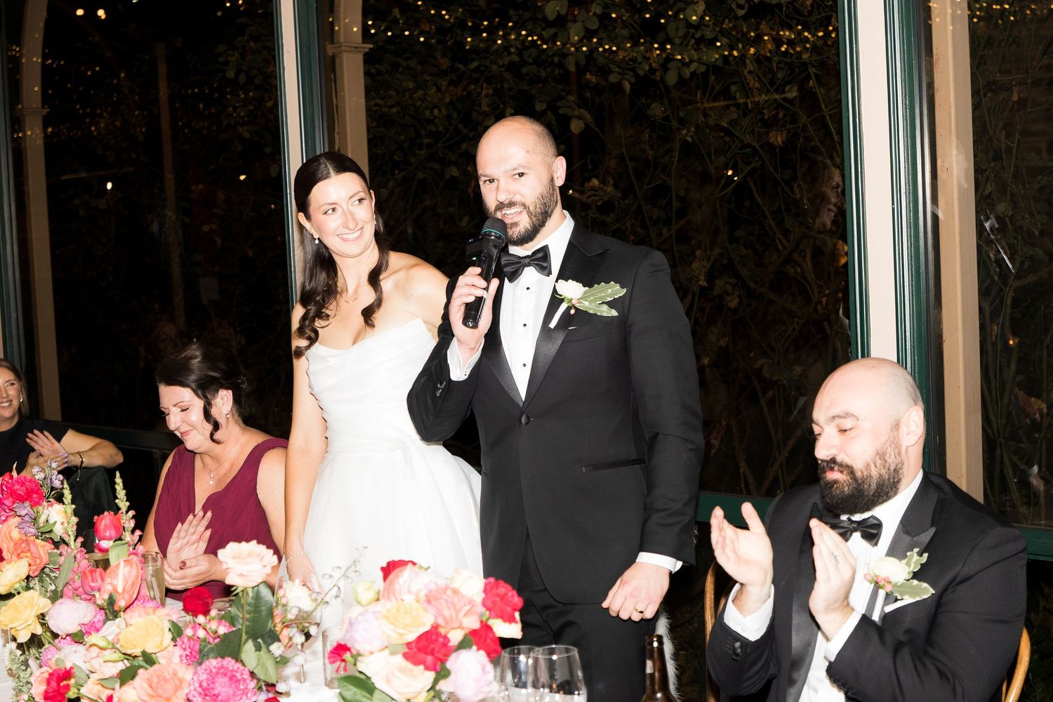 Bride and groom speak into microphone at wedding reception, surrounded by colorful floral arrangements and guests in formal attire.