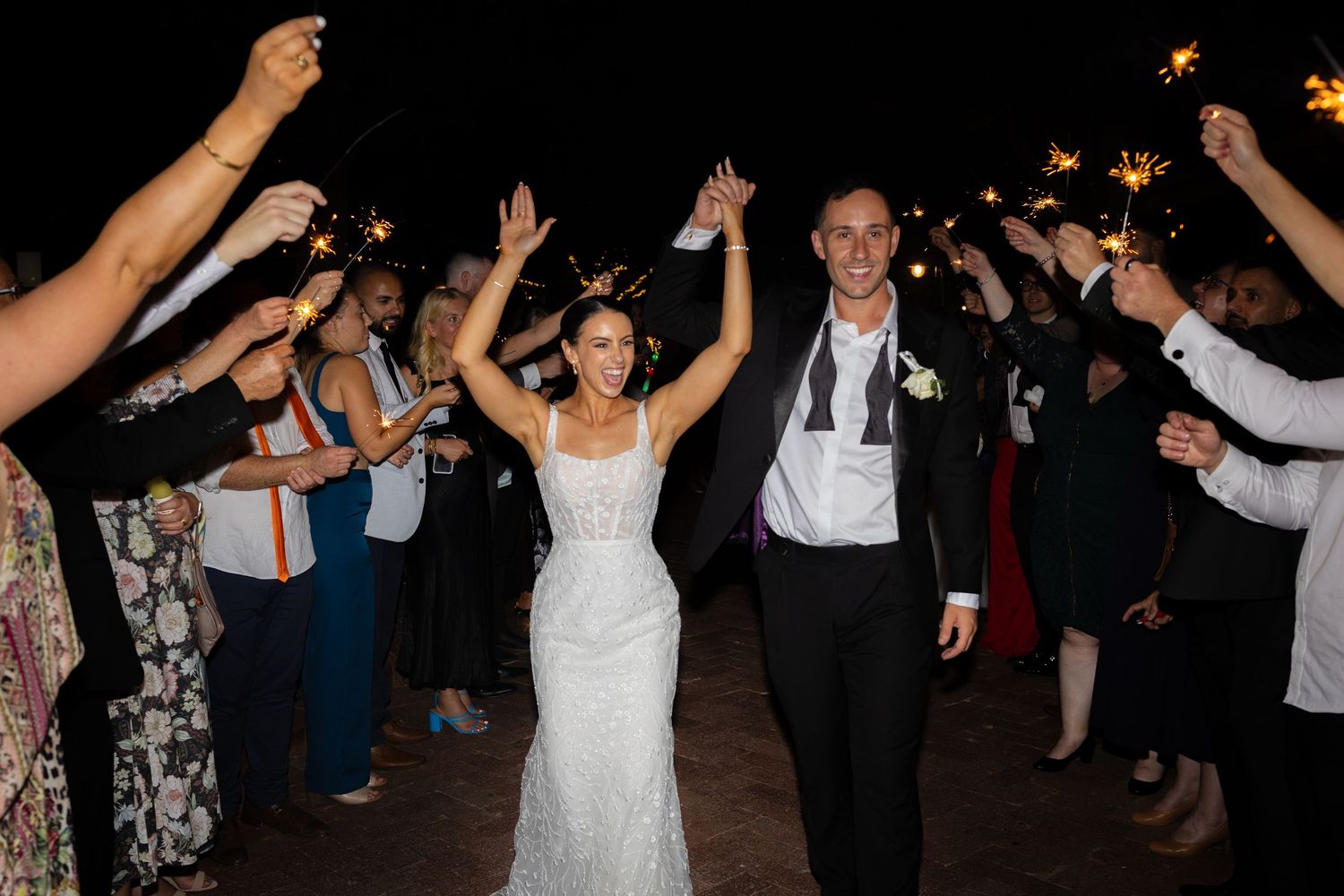 Newlyweds walk through a sparkler tunnel surrounded by cheering guests at their evening wedding reception.