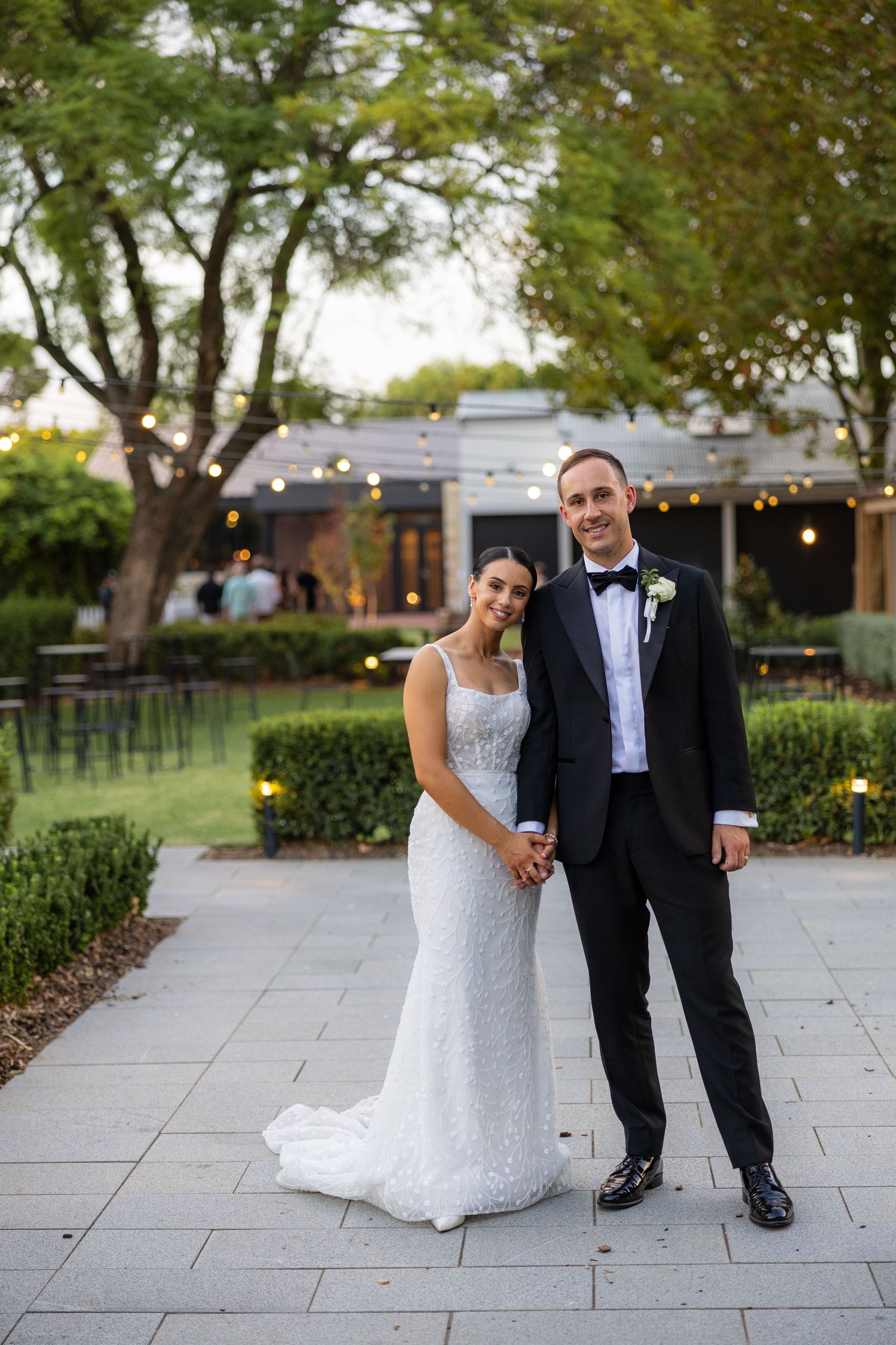 Bride in lace gown and groom in black tuxedo posing together in a garden venue at dusk.