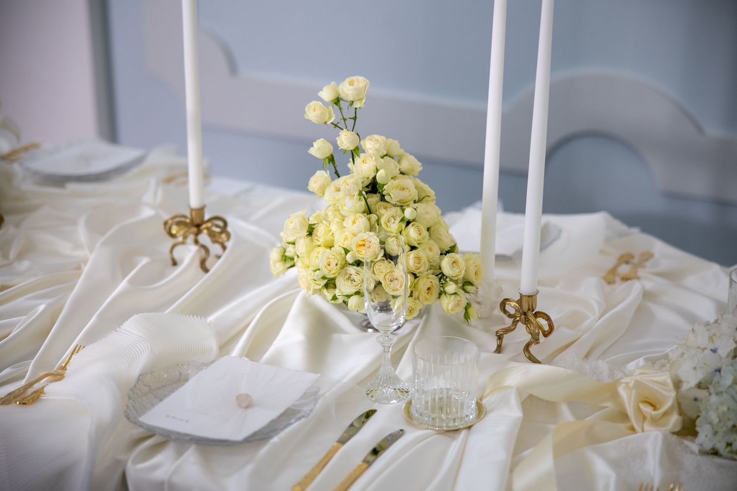Close-up of a wedding table with cream roses in a glass vase, white candles, and gold bow accents on ivory fabric.