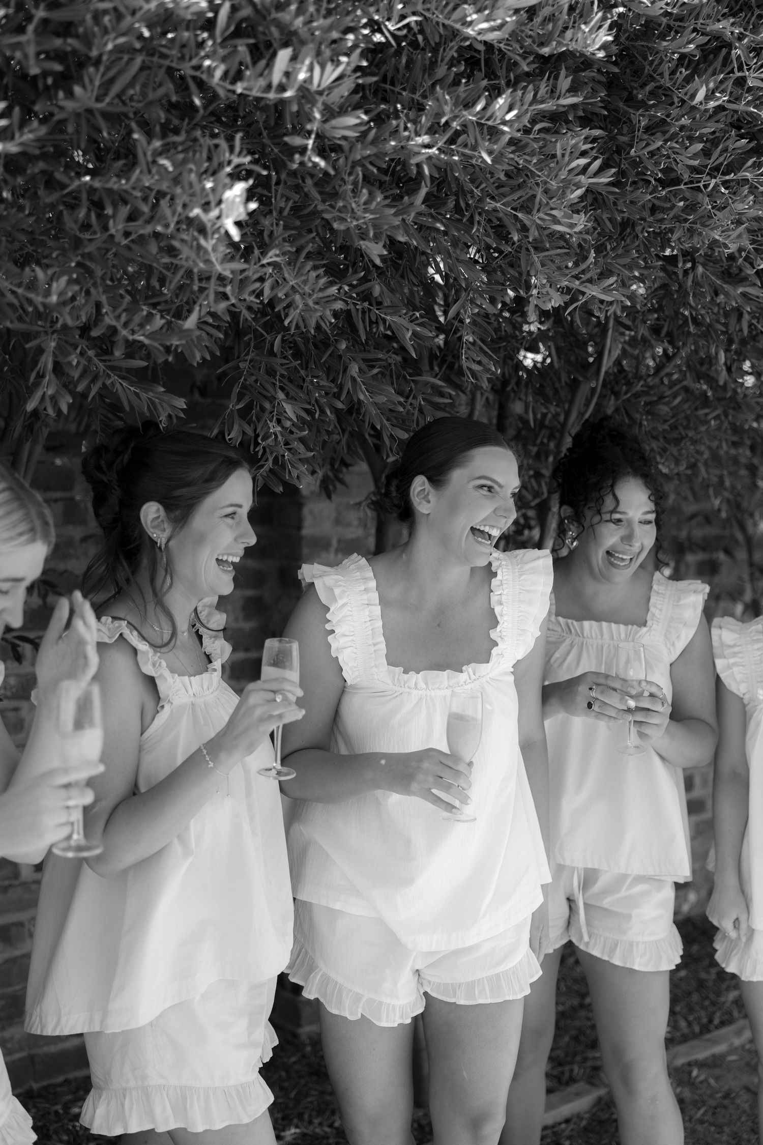 Black and white image of bridesmaids in white ruffled outfits laughing and holding champagne glasses.