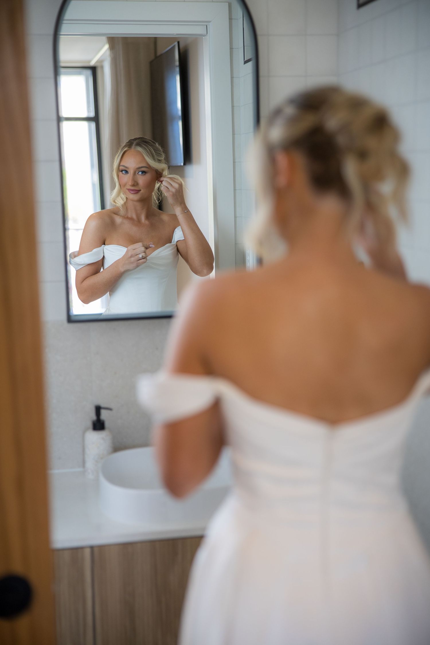 Blonde bride in off-shoulder white gown adjusting earring while looking in bathroom mirror on wedding day.