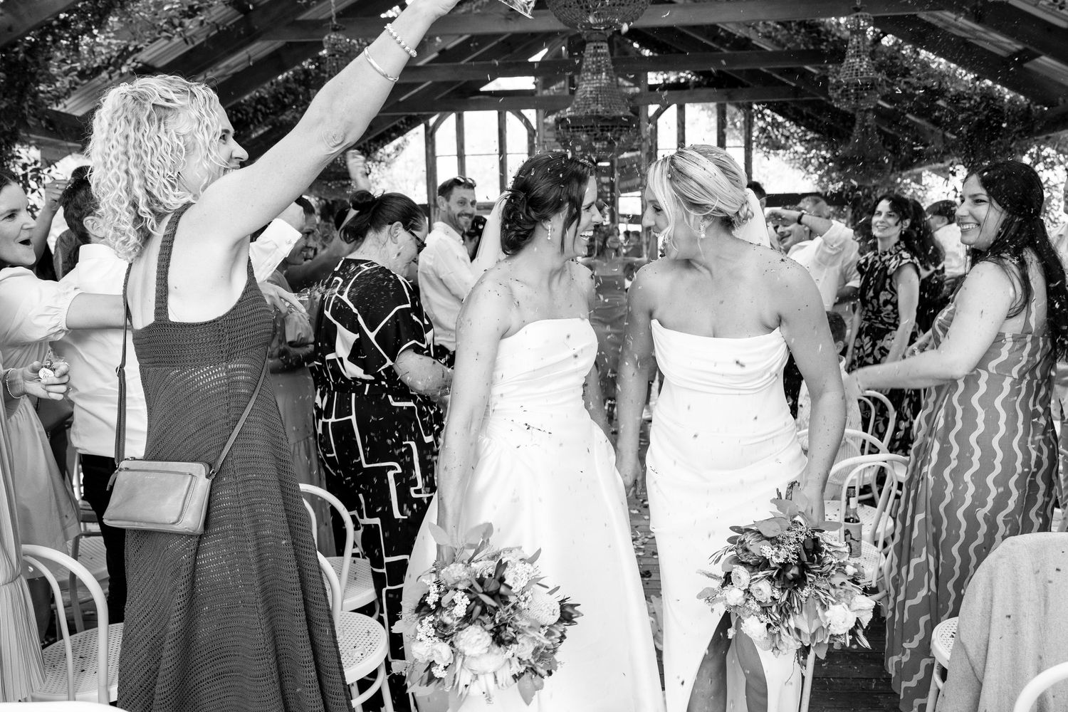 Black and white photo of two brides walking down the aisle surrounded by cheering wedding guests.