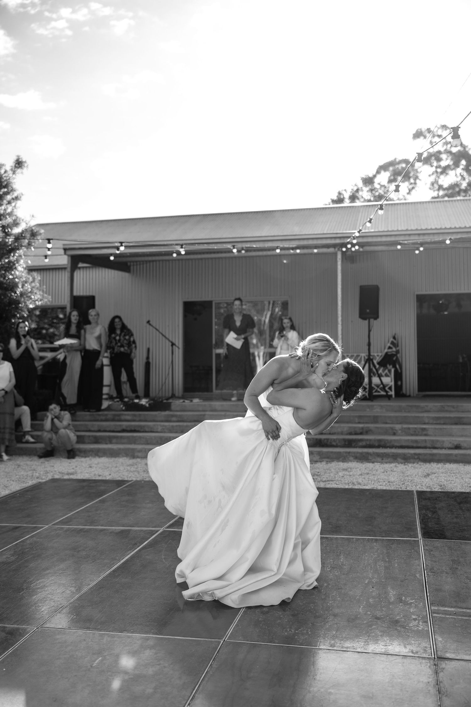 Black and white photo of two brides sharing a romantic dip kiss during their first dance outdoors.