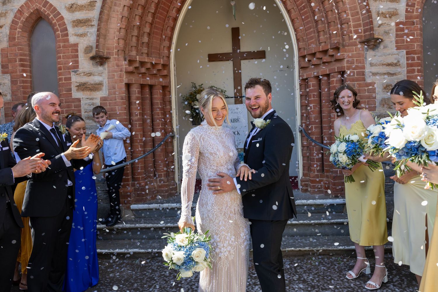 Bride and groom walk out of a brick church entrance as guests toss confetti in joyful celebration.